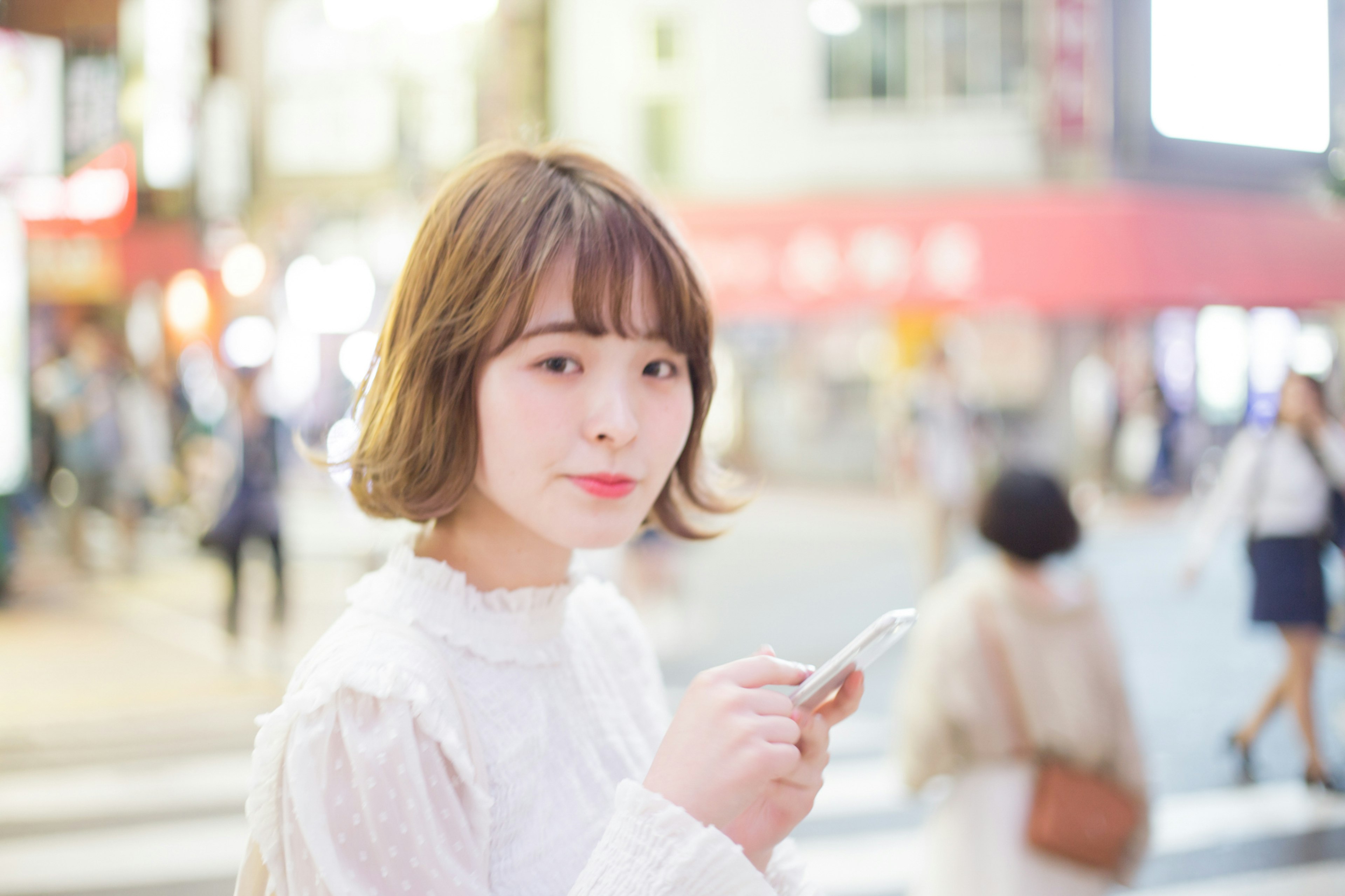 Porträt einer Frau, die an einer Straßenecke ein Smartphone hält Sie trägt ein weißes Outfit mit verschwommenen Menschen im Hintergrund