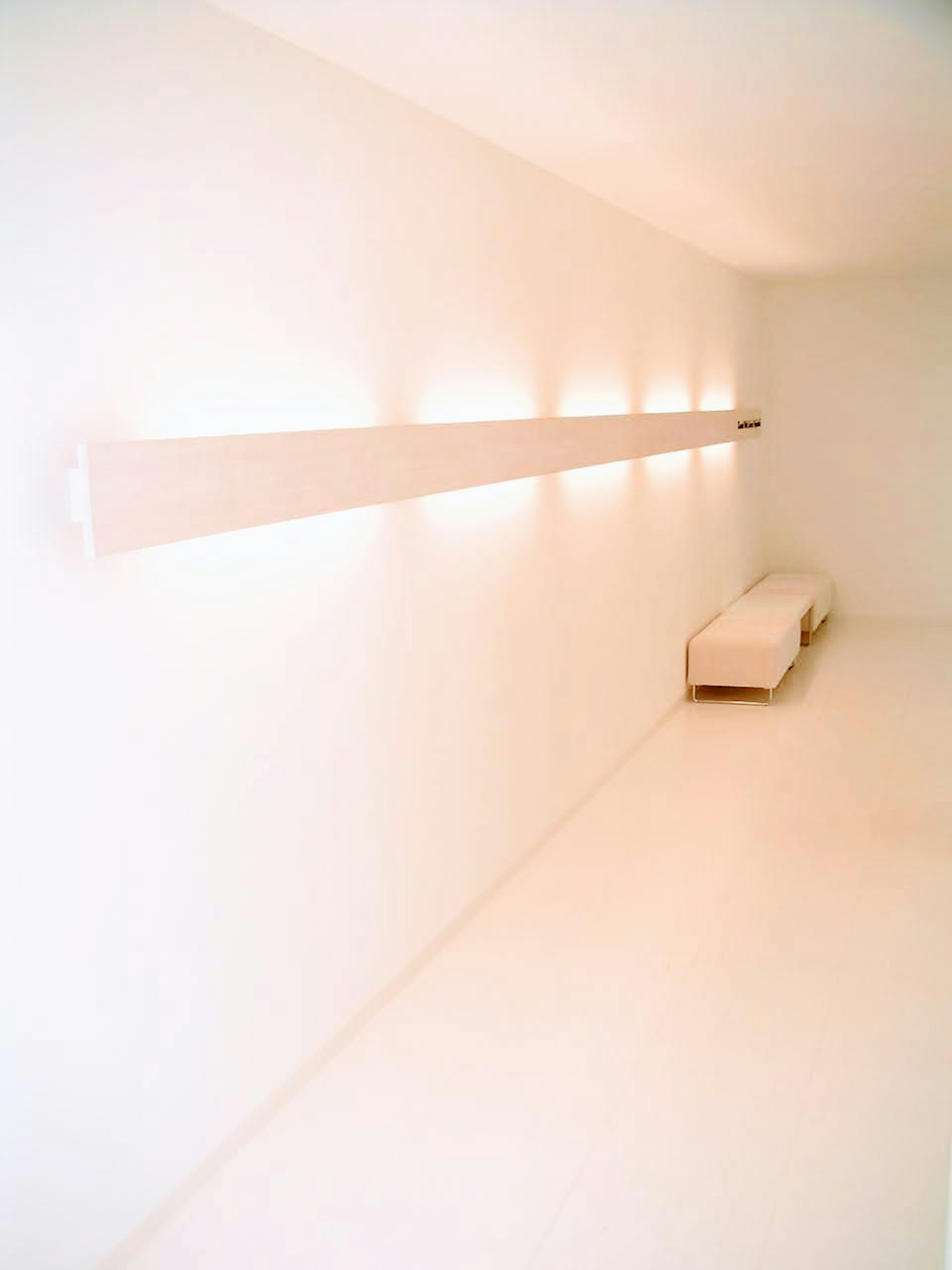 A minimalistic room with white walls and a long light fixture with a white sofa