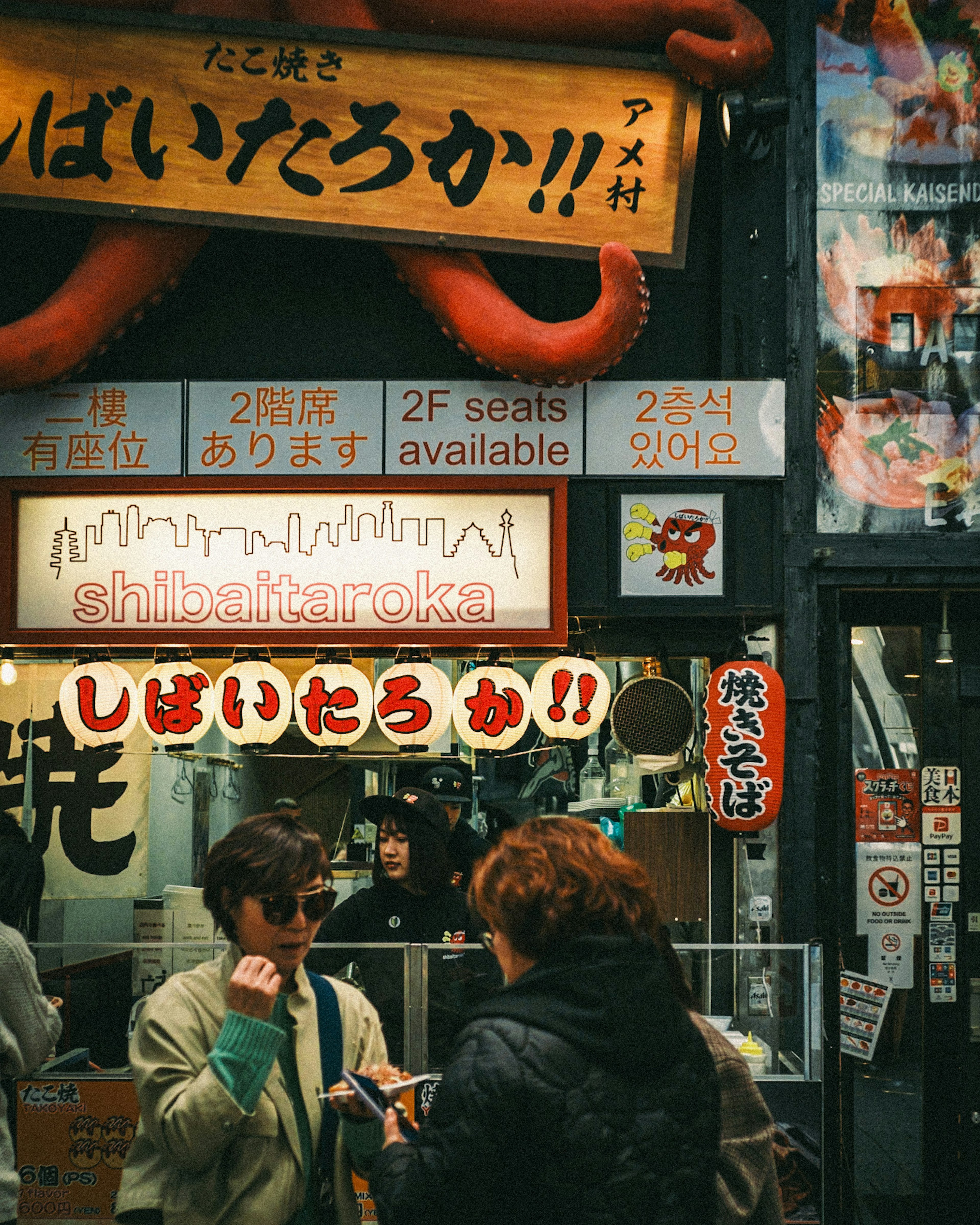 Fachada de una tienda de comida callejera japonesa con un llamativo cartel de pulpo y personas