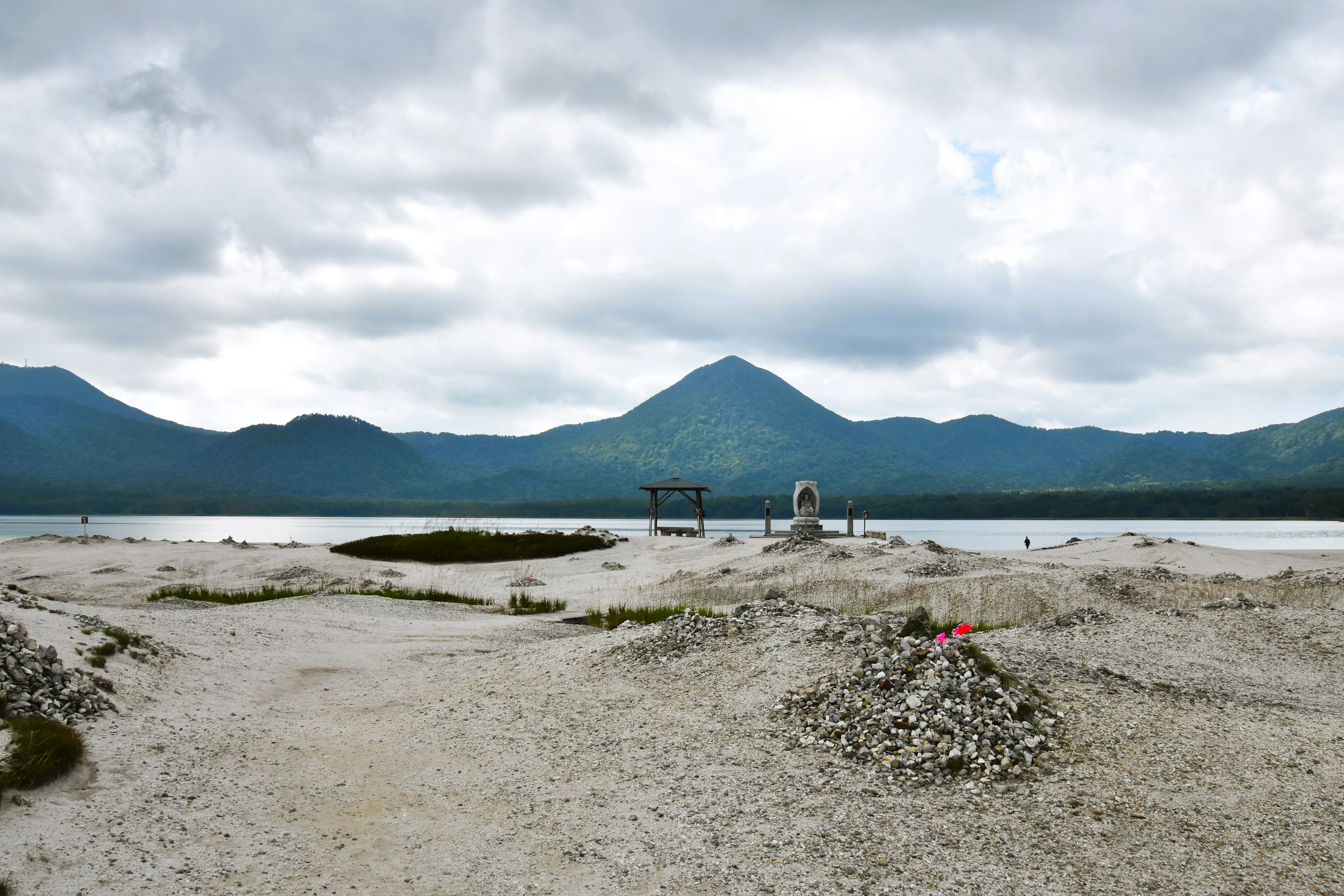 Expansive landscape of gray rocks and sand with mountains and cloudy sky in the background near a lake