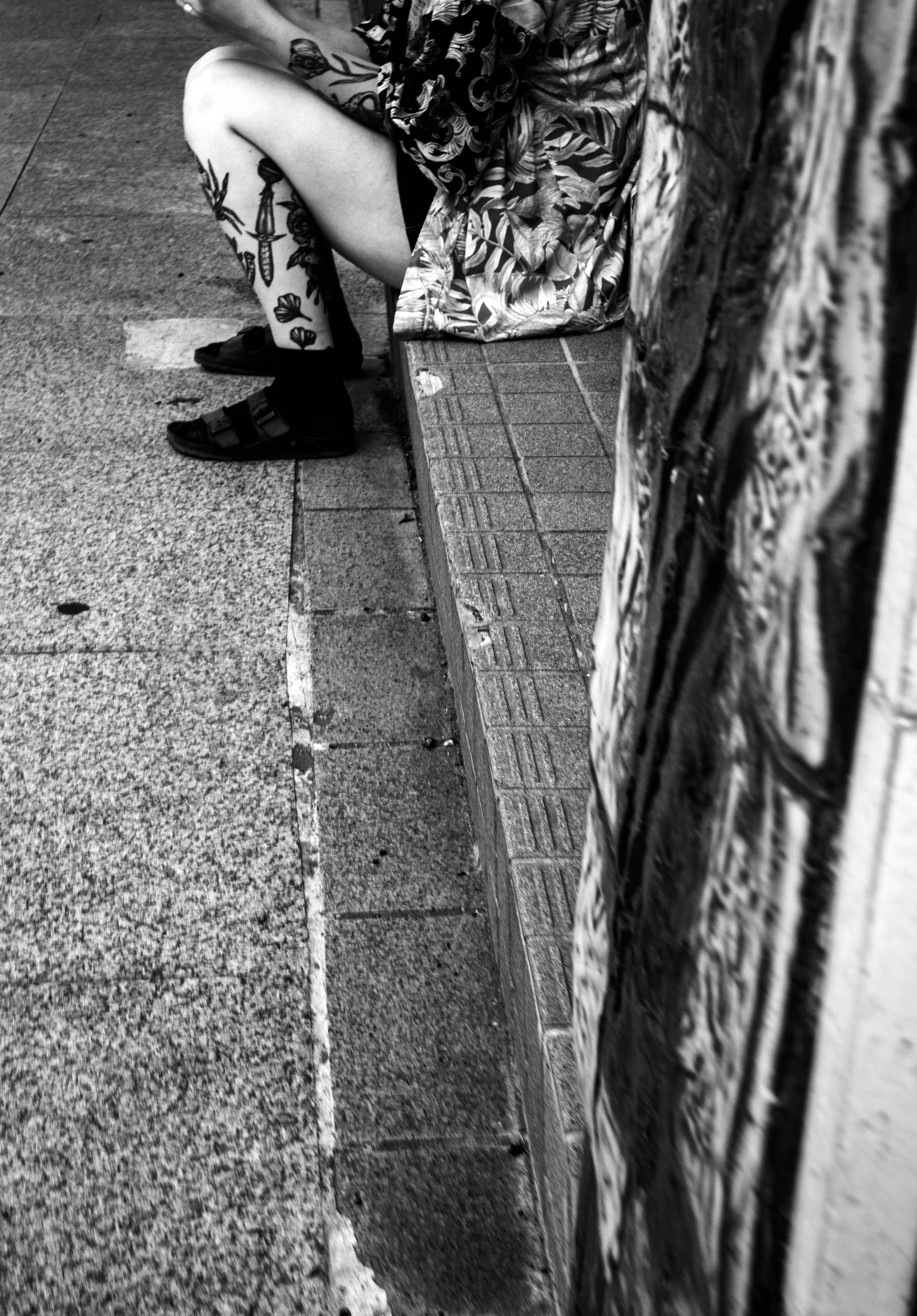 Black and white image featuring a person sitting on a bench wearing a floral dress and showing their legs