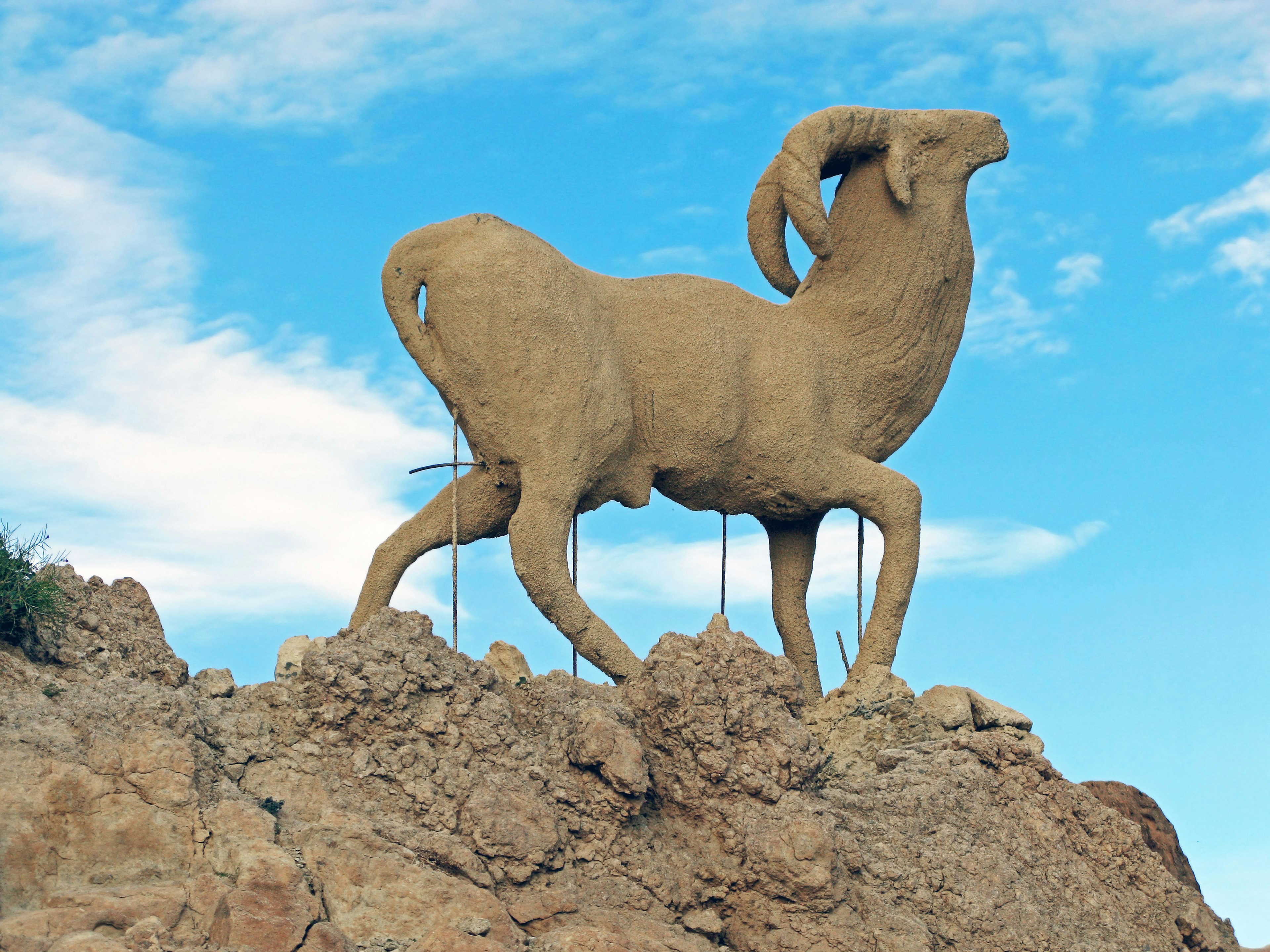 Grande scultura di un ariete in piedi su una roccia con uno sfondo di cielo blu e nuvole bianche