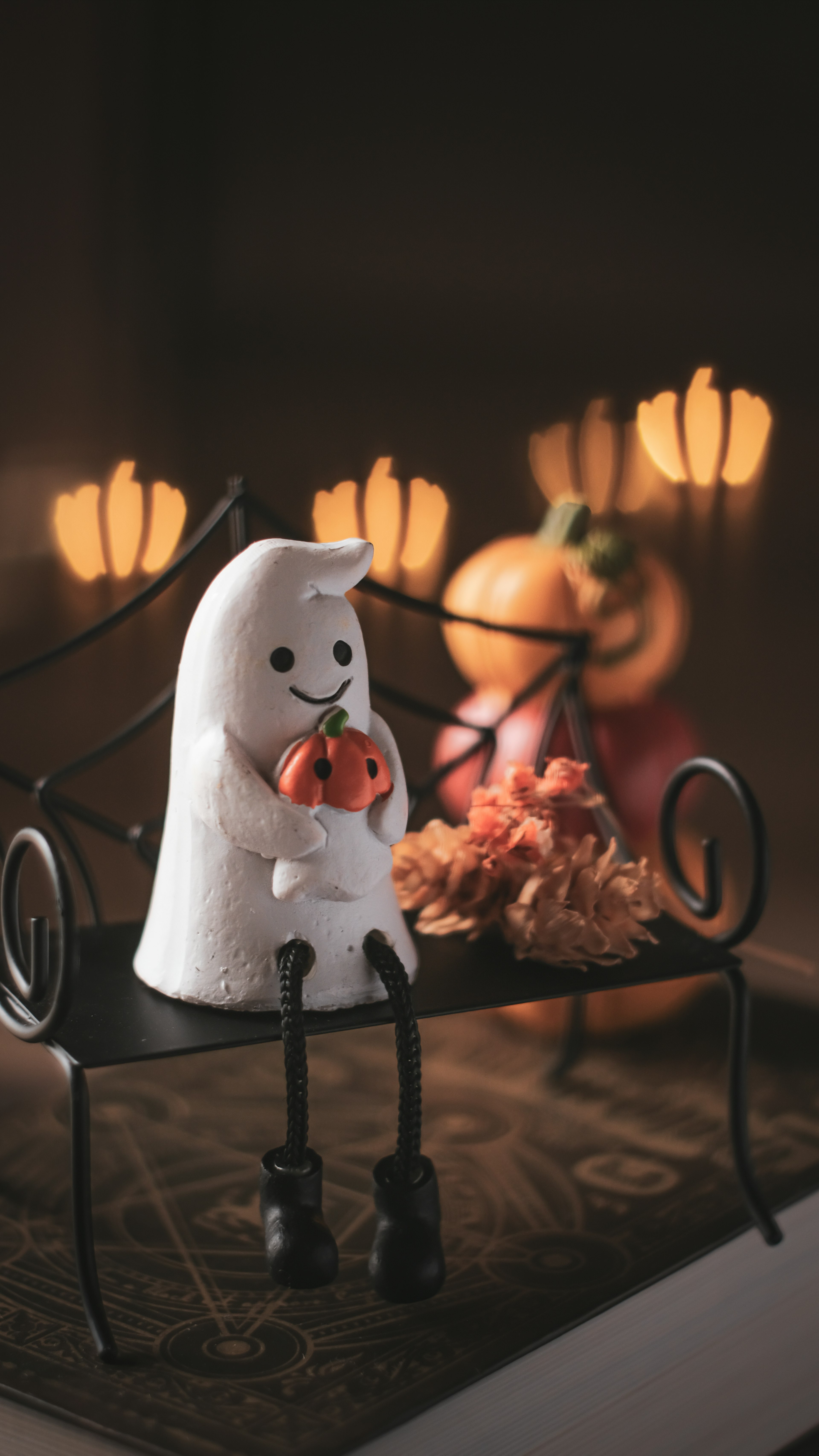 A white ghost figurine sitting on a small bench with orange pumpkins and flowers in the background