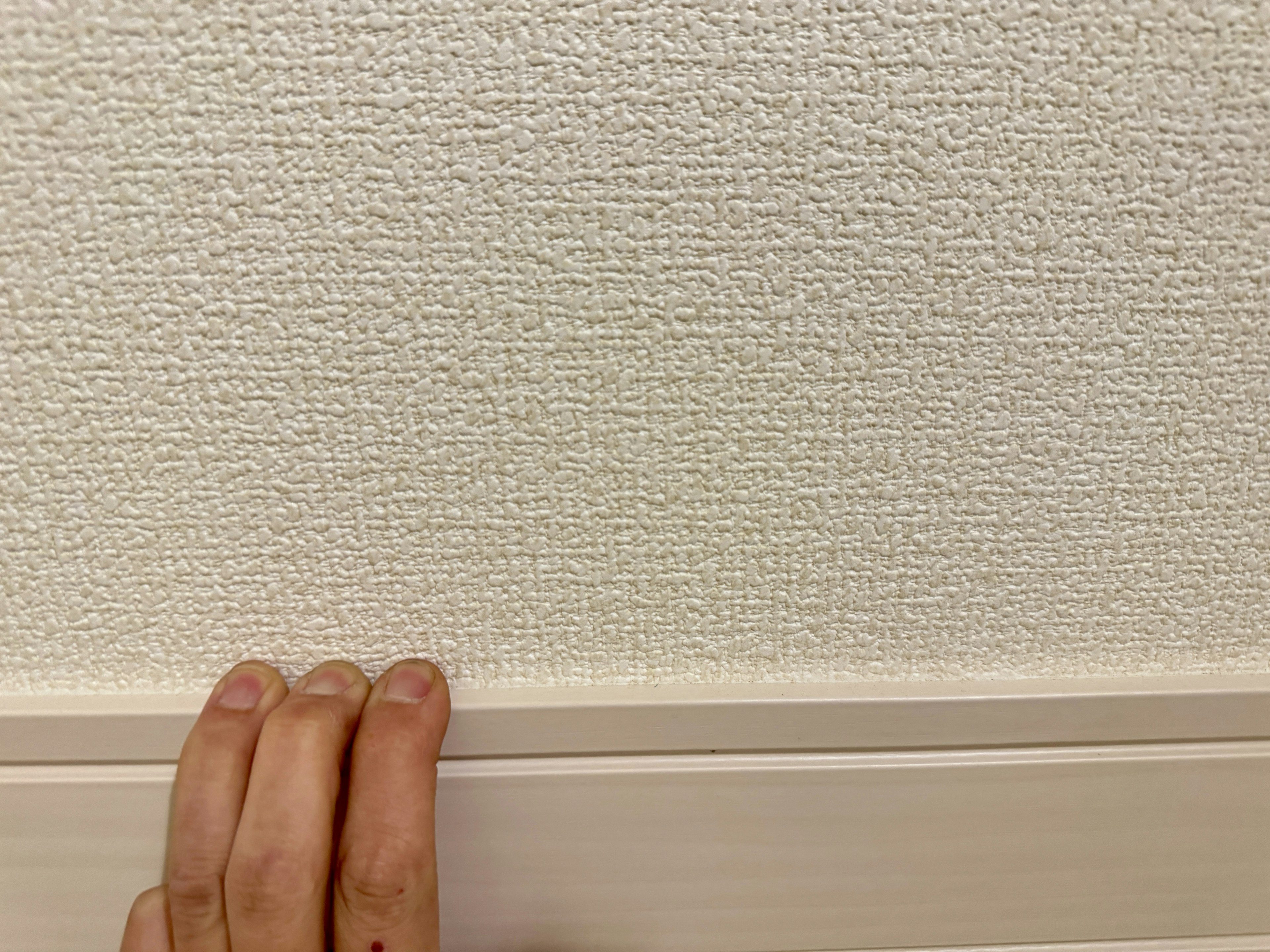 A hand touching a textured white wall with wooden molding at the bottom