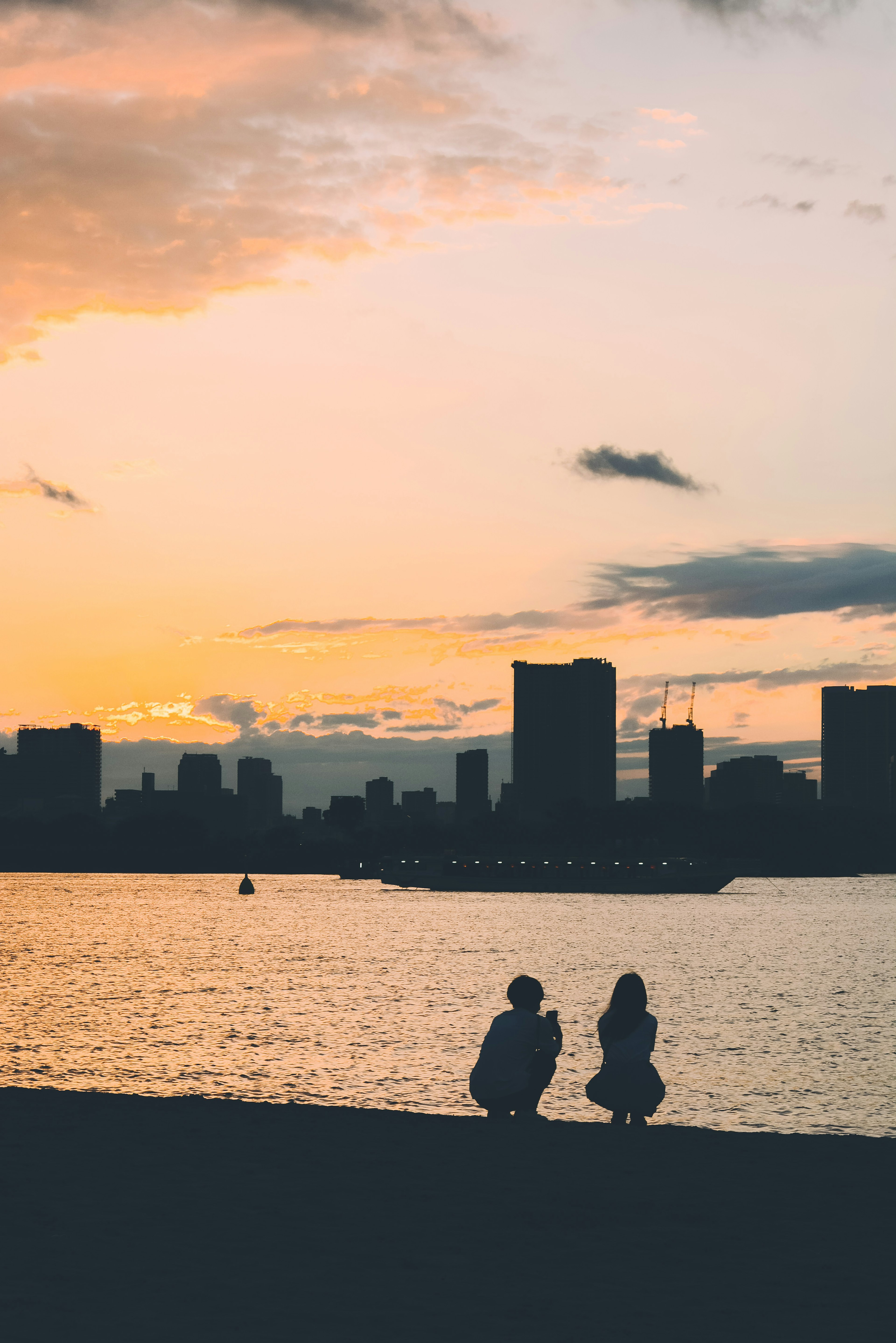 夕日を背景にした海辺で座っている二人の人物と都市のシルエット