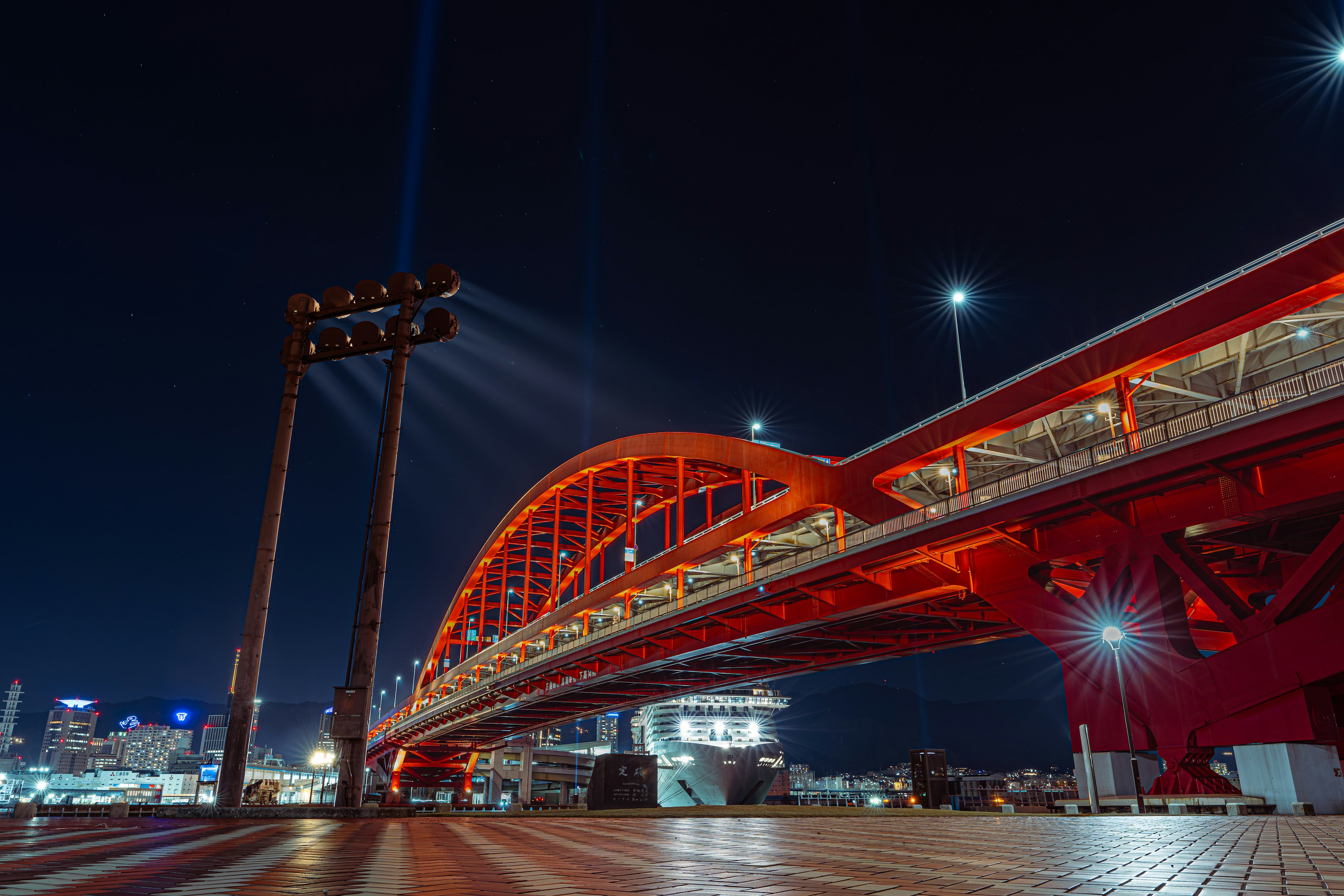 Jembatan lengkung merah yang diterangi di malam hari dengan lampu kota di latar belakang