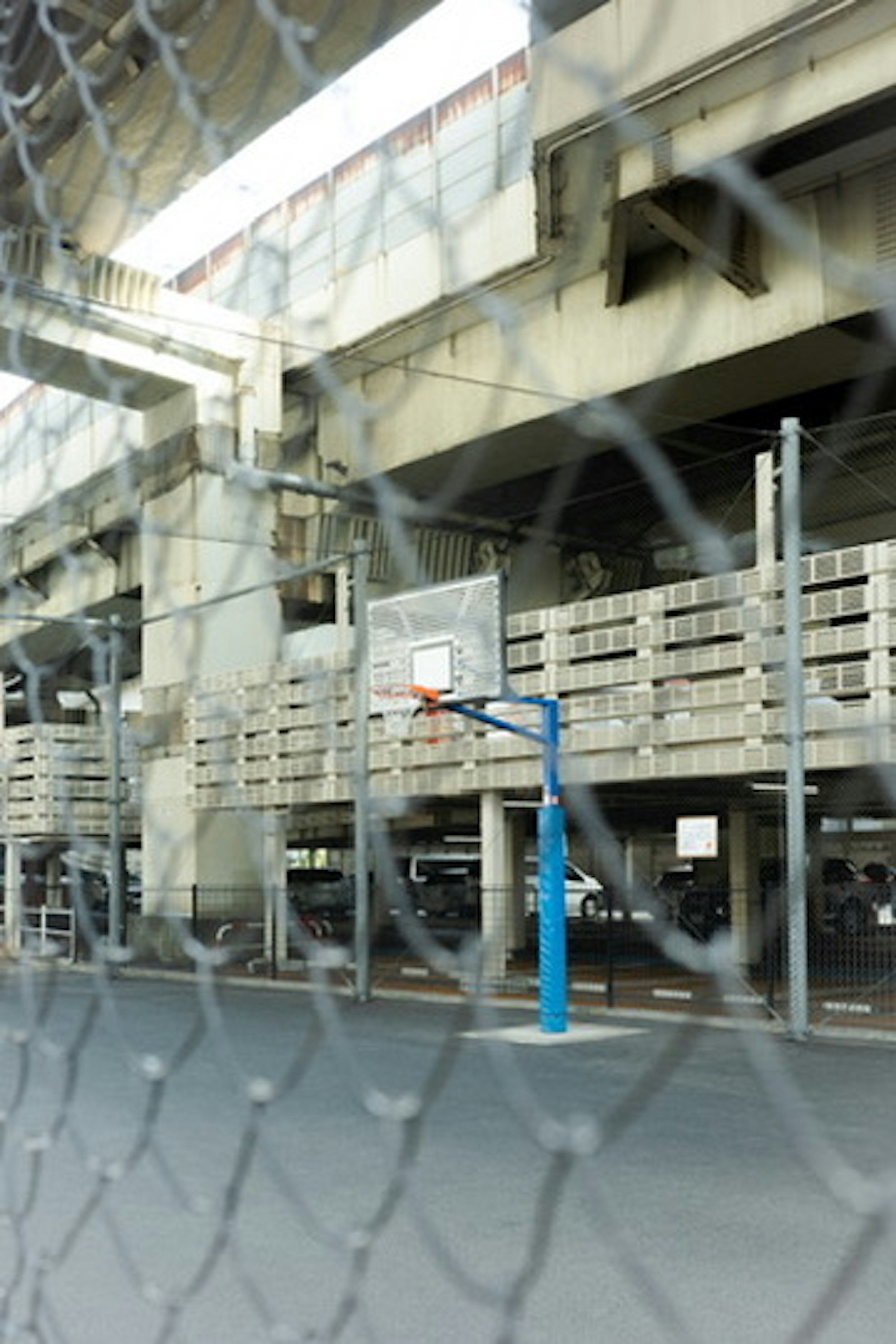 Basketball hoop with blue post under an overpass surrounded by a chain-link fence