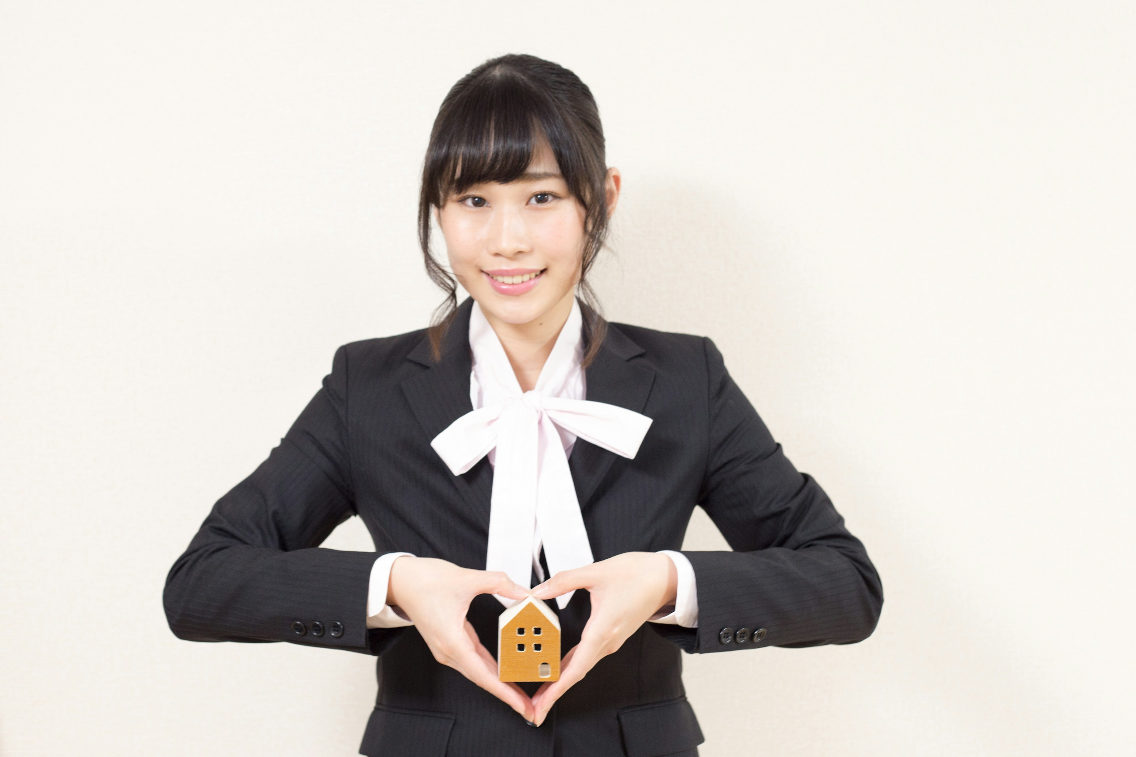 A woman in a suit forms a heart shape with her hands while holding a small house model