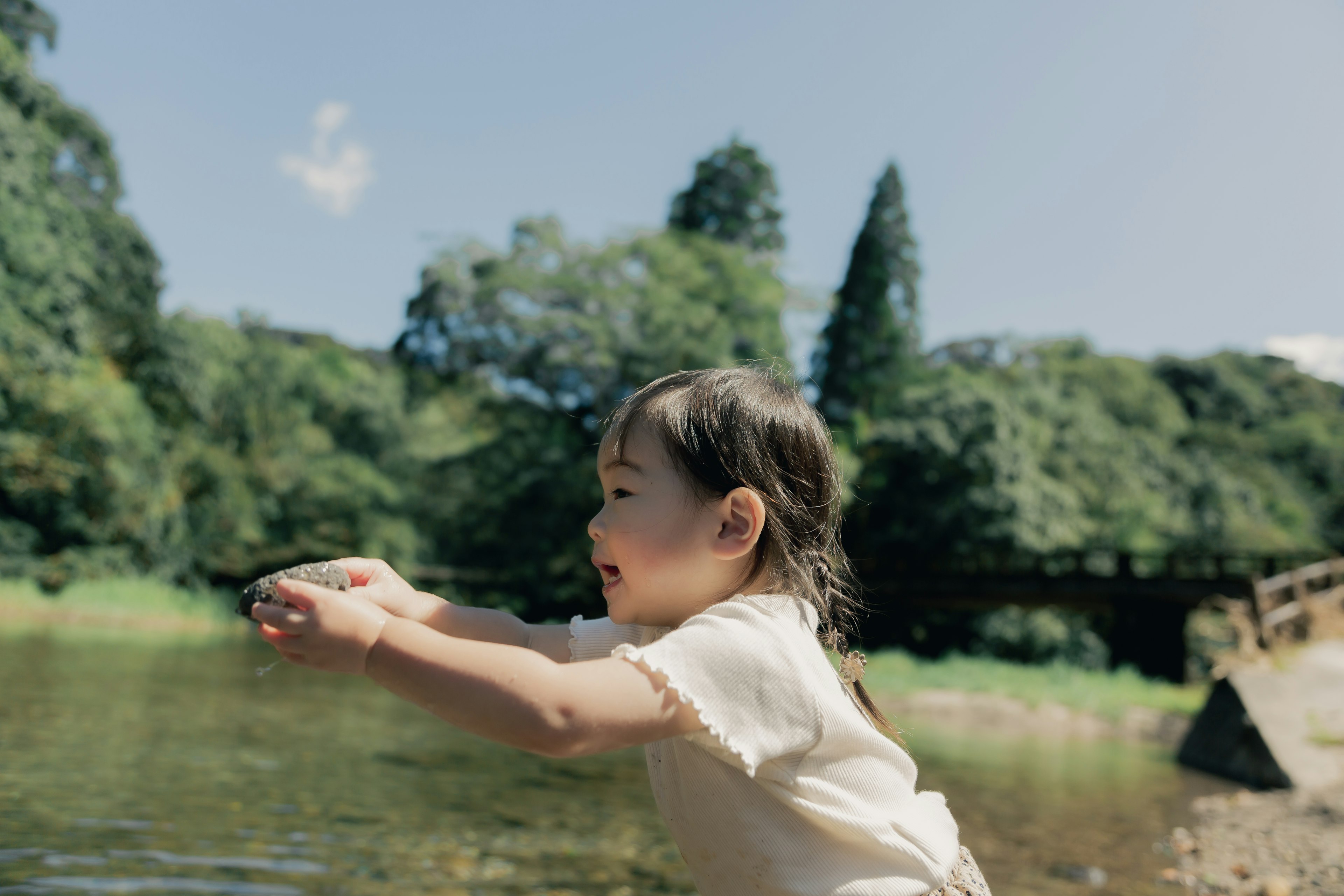 Ein junges Mädchen spielt am Fluss umgeben von grünen Bäumen und blauem Himmel