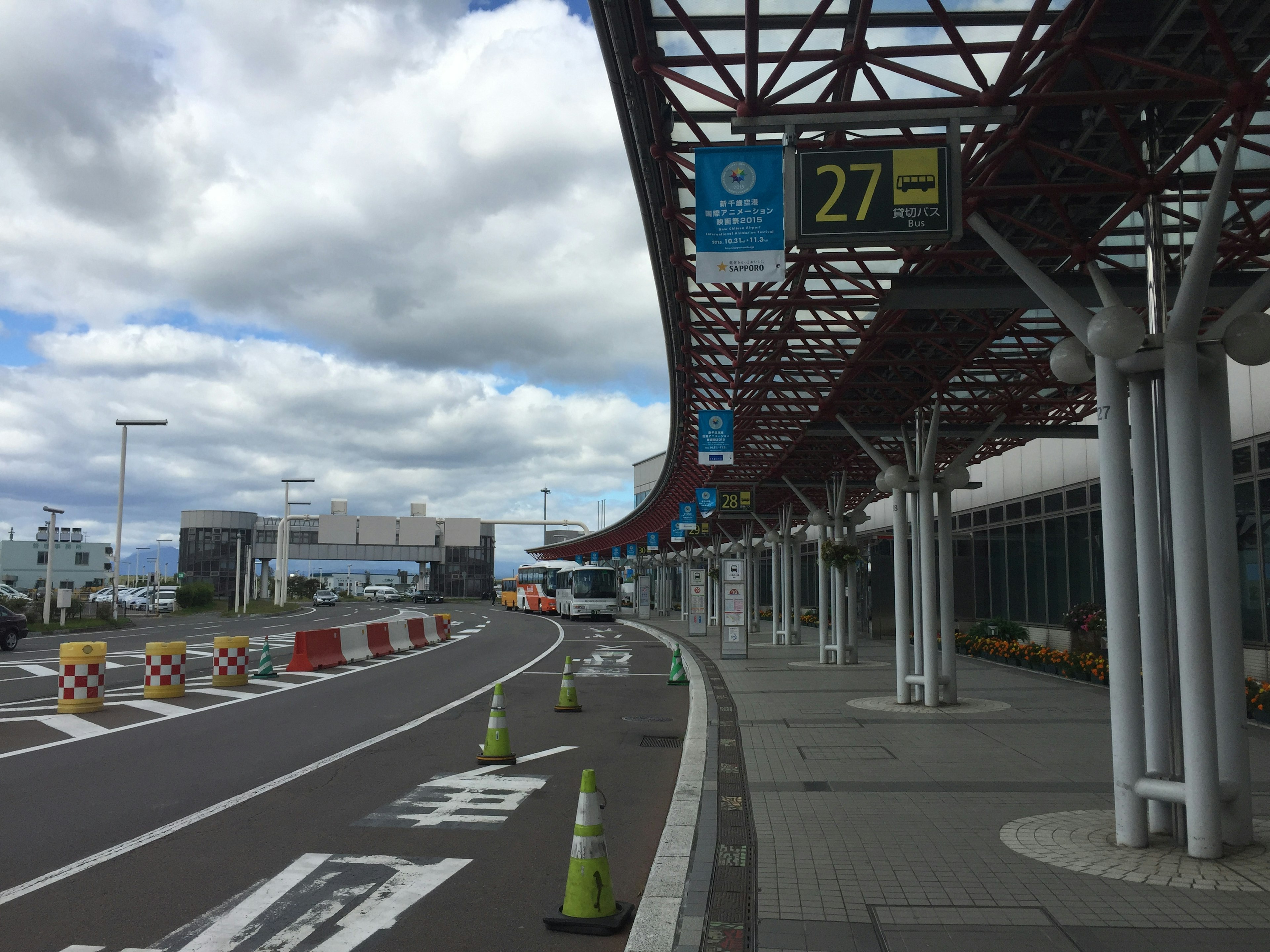 Exterior view of an airport departure area with blue sky and clouds showing sign 27 and traffic cones placed along the road