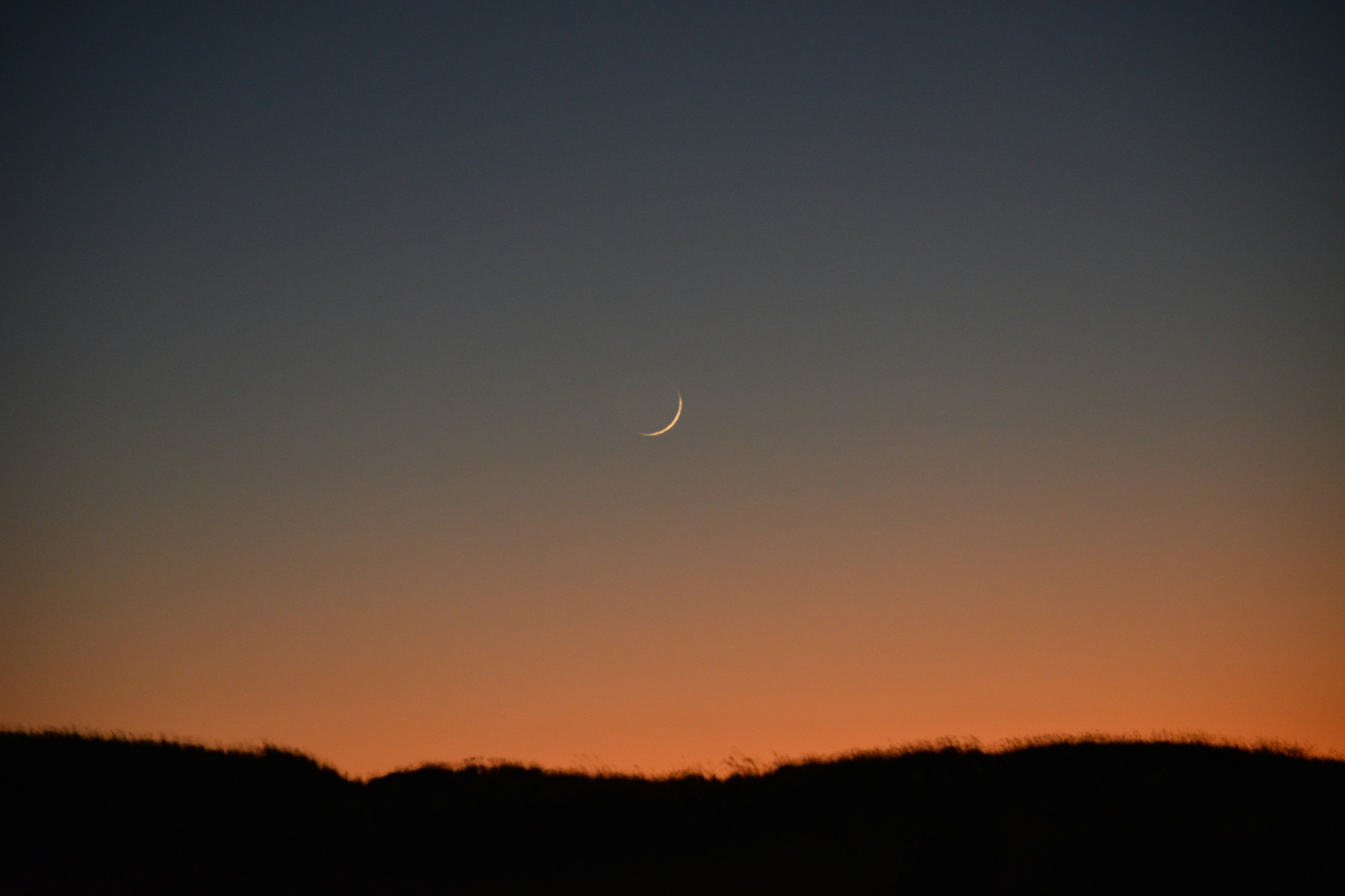 Luna crescente sottile nel cielo crepuscolare con silhouette di montagna arancione