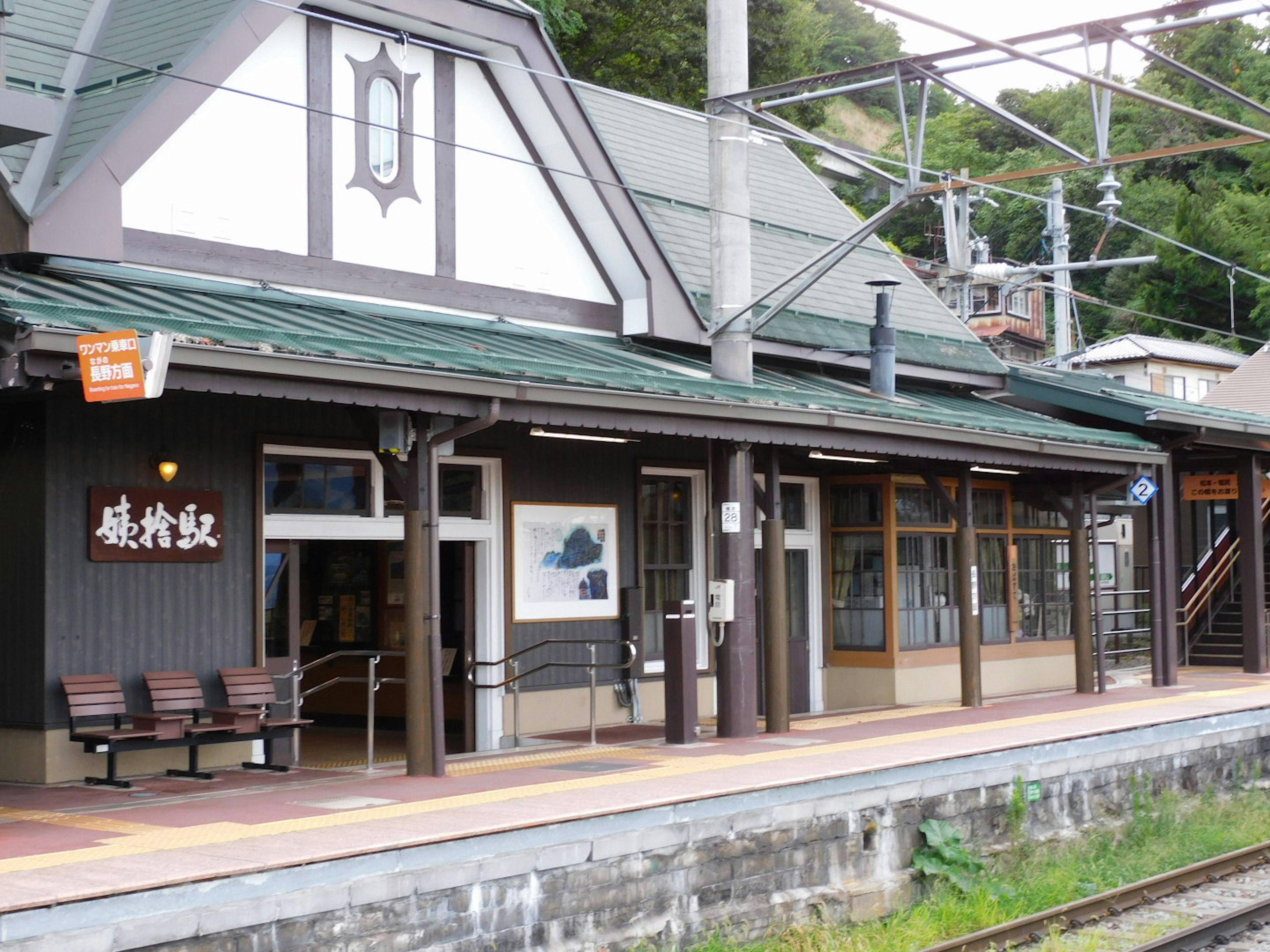 ภายนอกสถานีที่มีหลังคาสีเขียวและพื้นที่รอรางรถไฟและม้านั่งที่มองเห็นได้