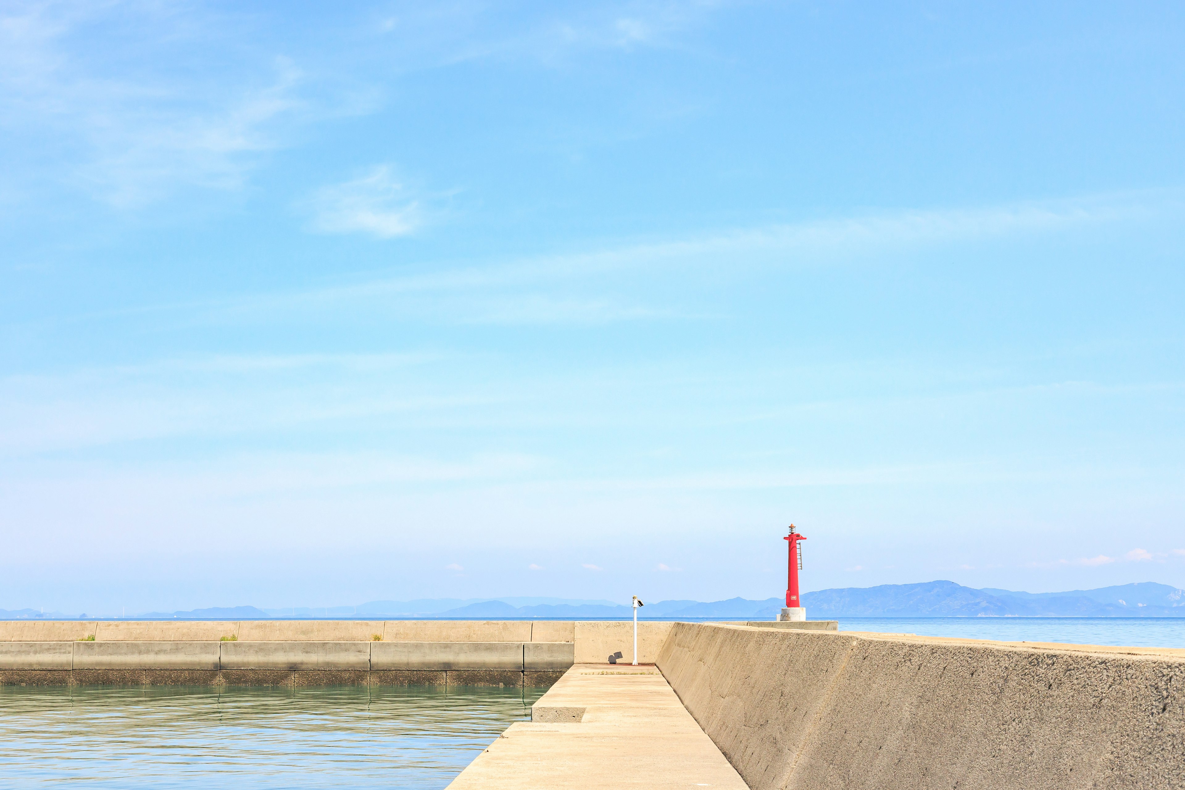 Ein Wellenbrecher mit einem roten Leuchtturm vor blauem Himmel und ruhiger See