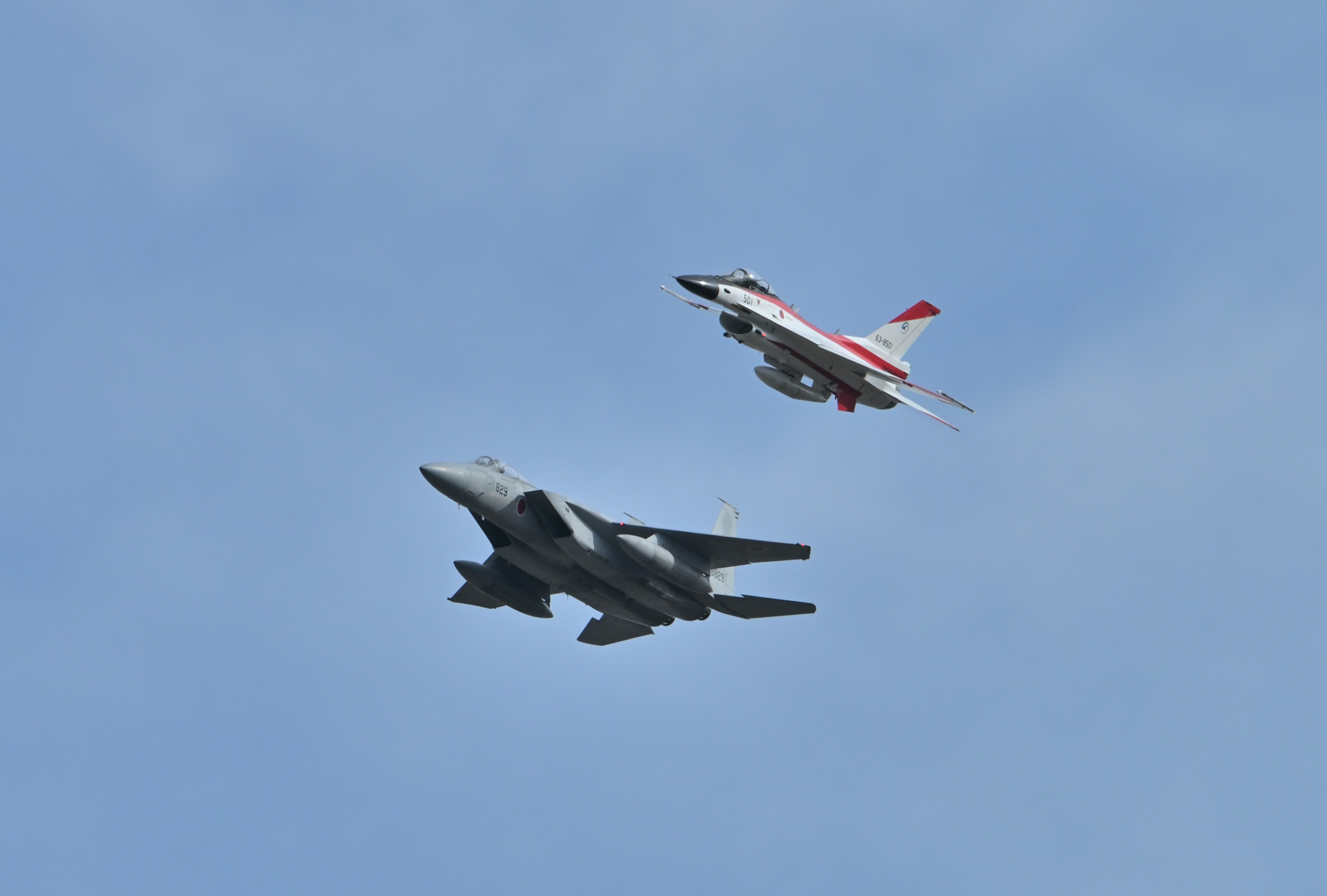 Two fighter jets flying in the sky one with a dark body and the other with a white and red body