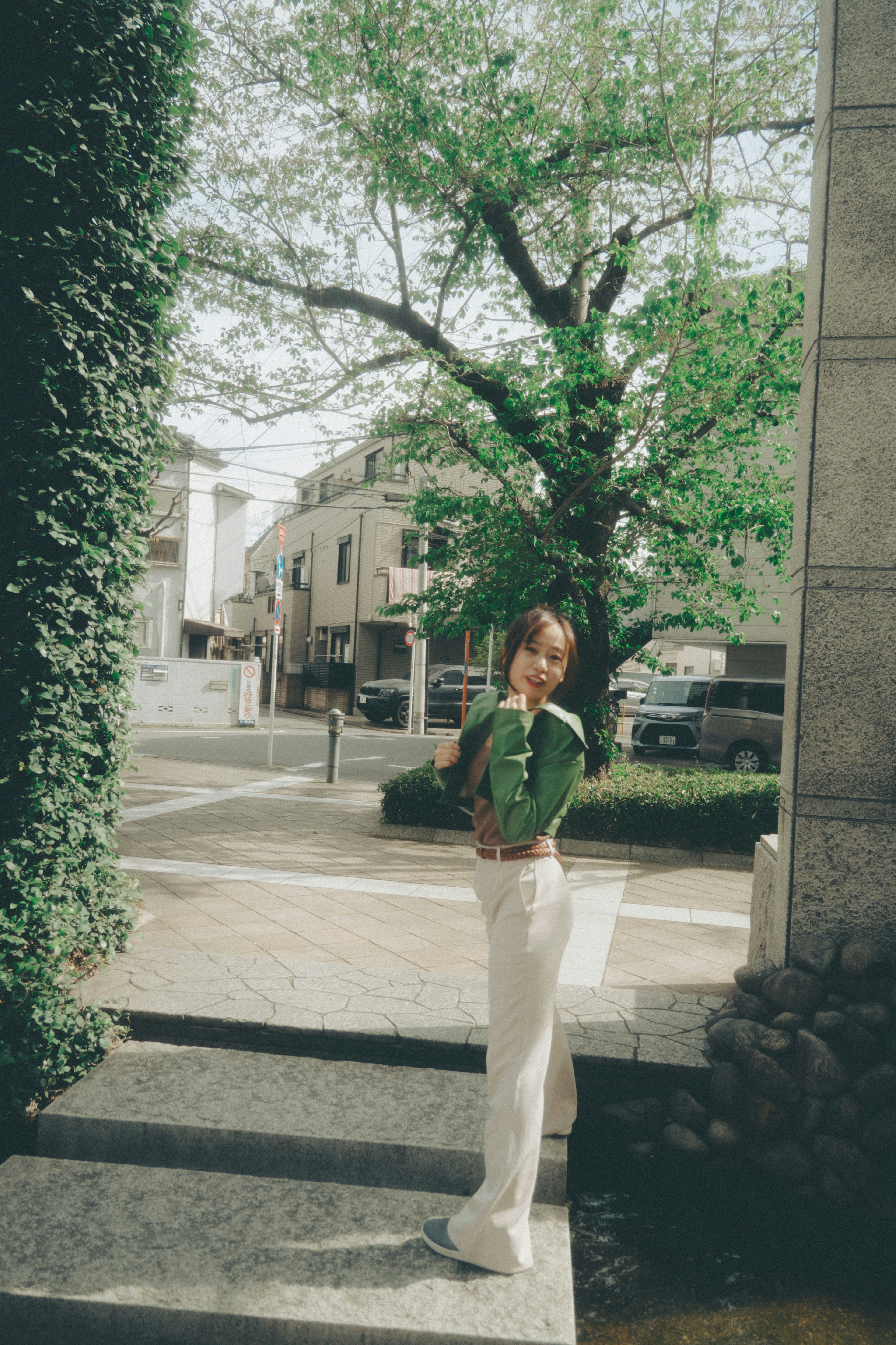 A woman standing in front of a green tree wearing light-colored pants and a green top