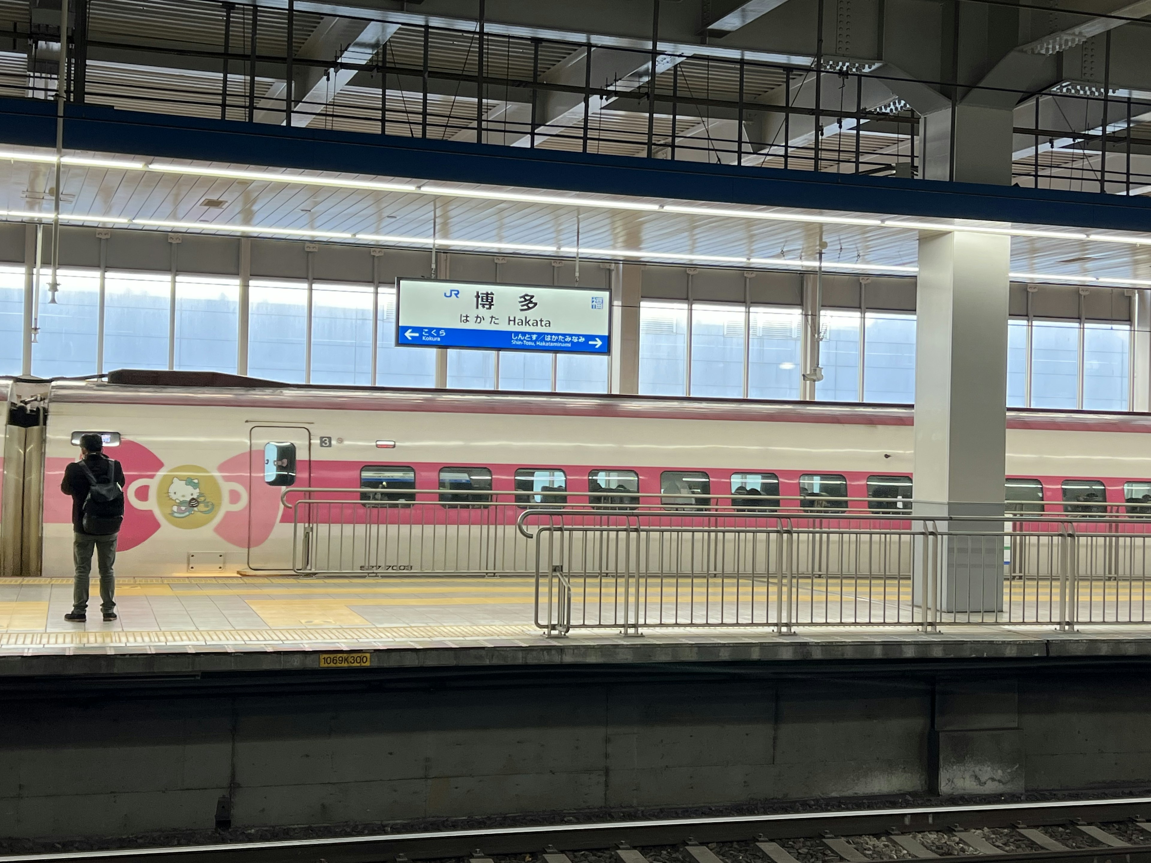 駅のプラットフォームに立つ人物とピンクの電車が見える風景