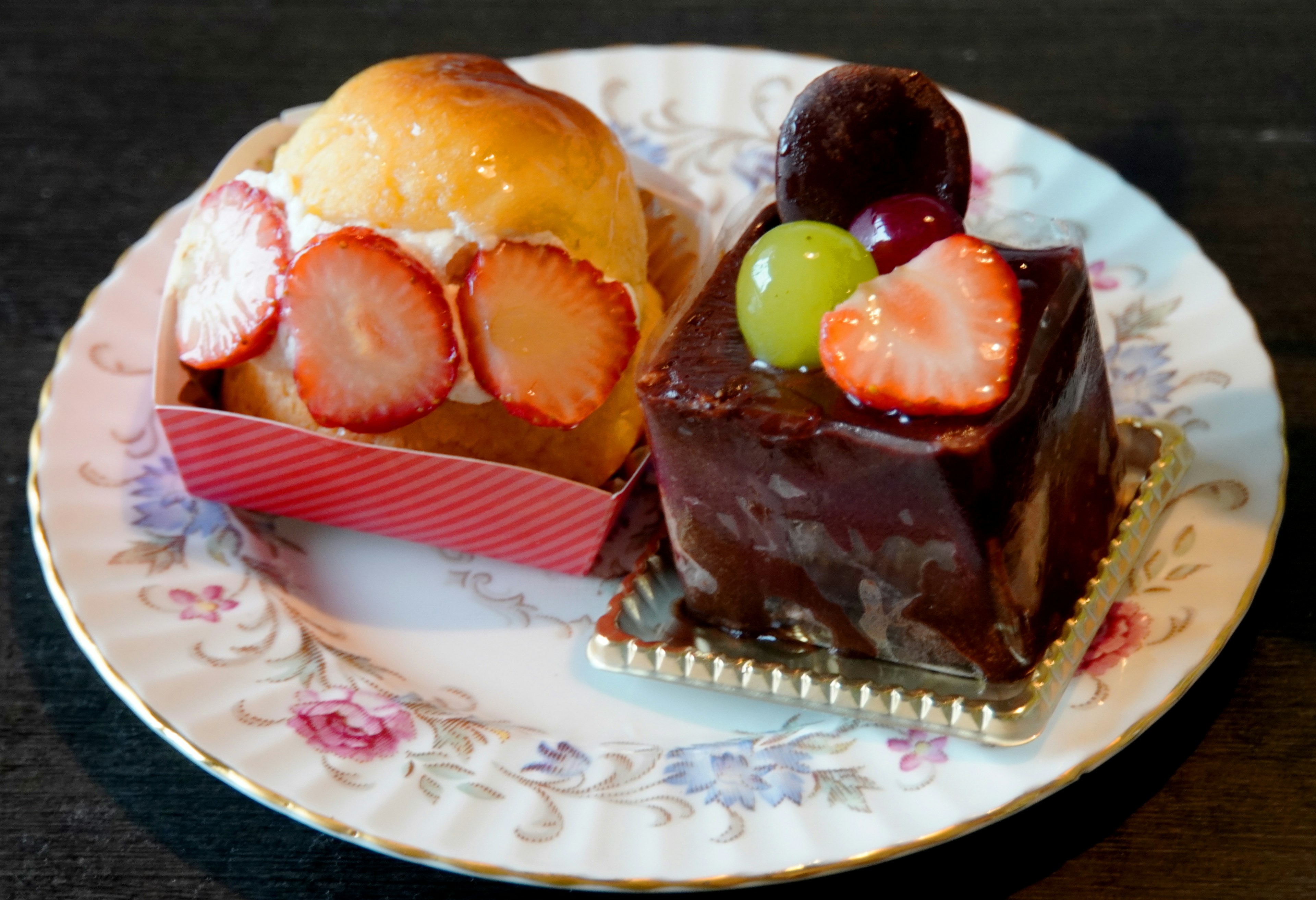 Sweet roll topped with strawberries and cream alongside a chocolate cake with fruit