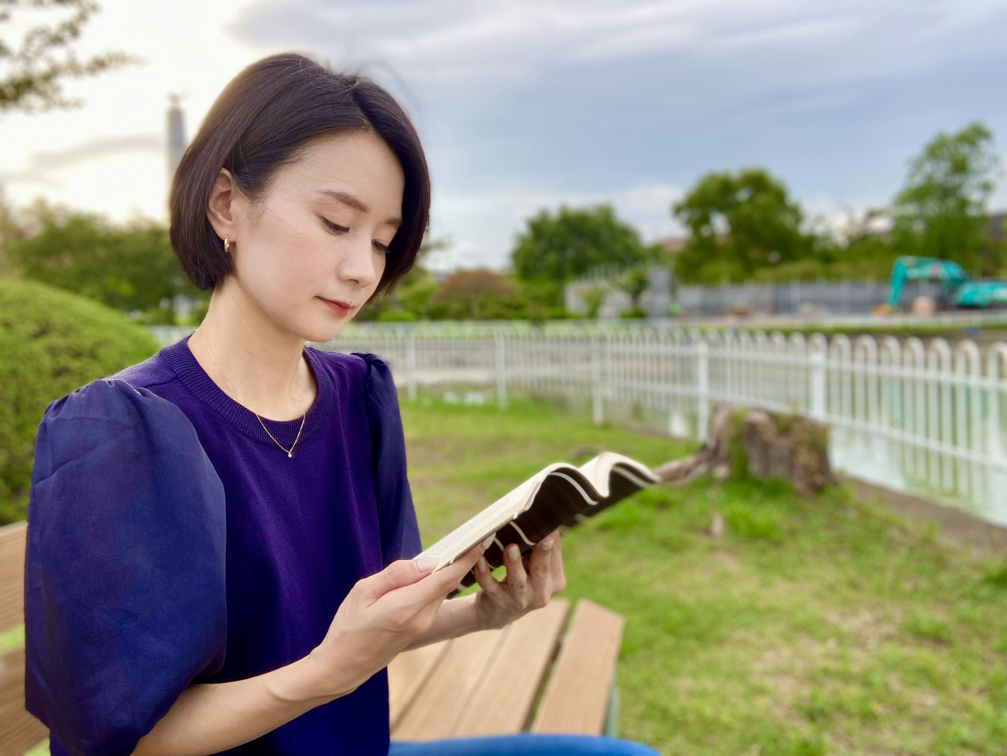 Eine Frau liest ein Buch in einem Park mit Grünflächen und einem weißen Zaun im Hintergrund