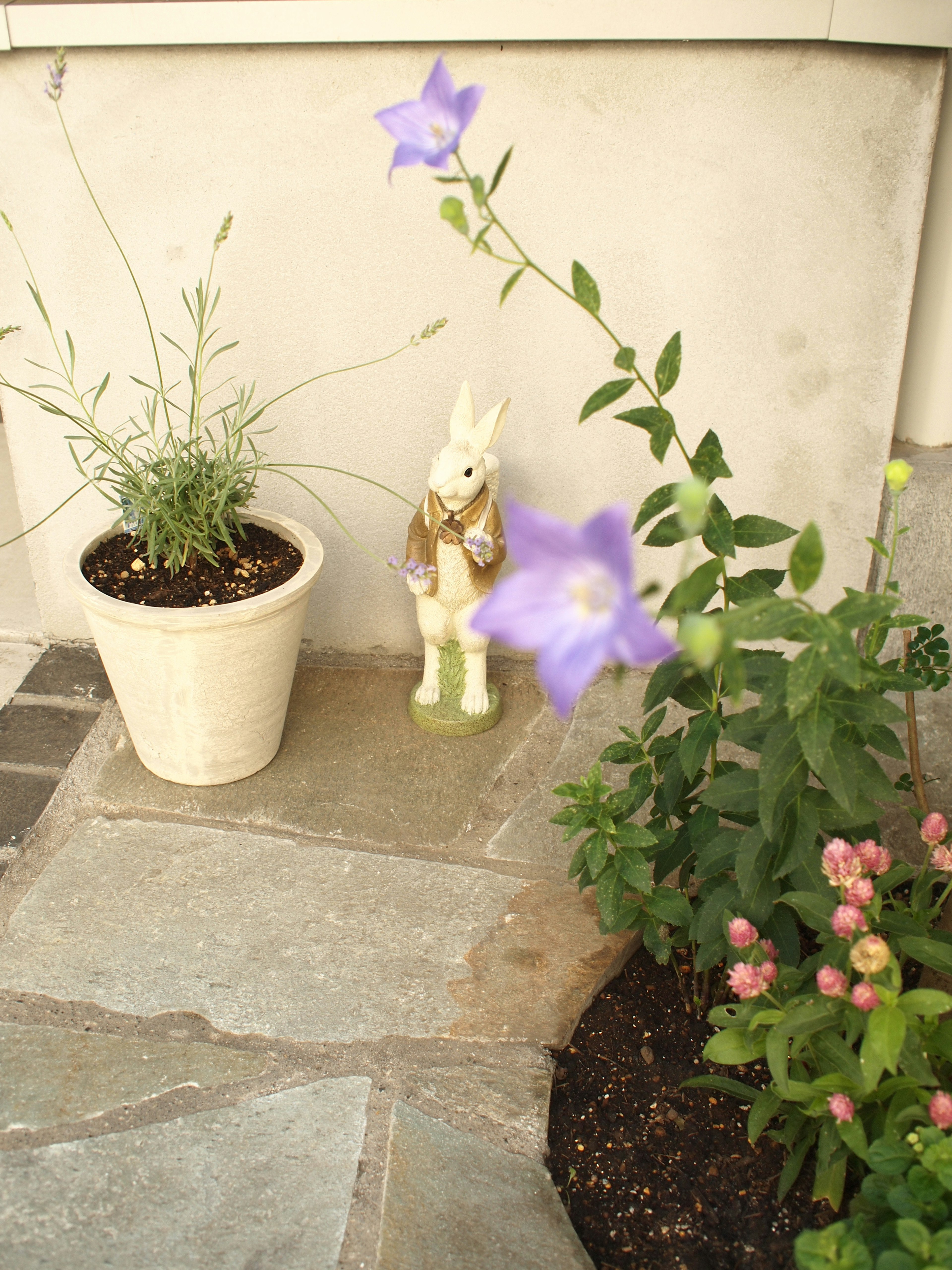 A decorative rabbit figurine standing near a white pot with herbs and purple flowers in a garden corner