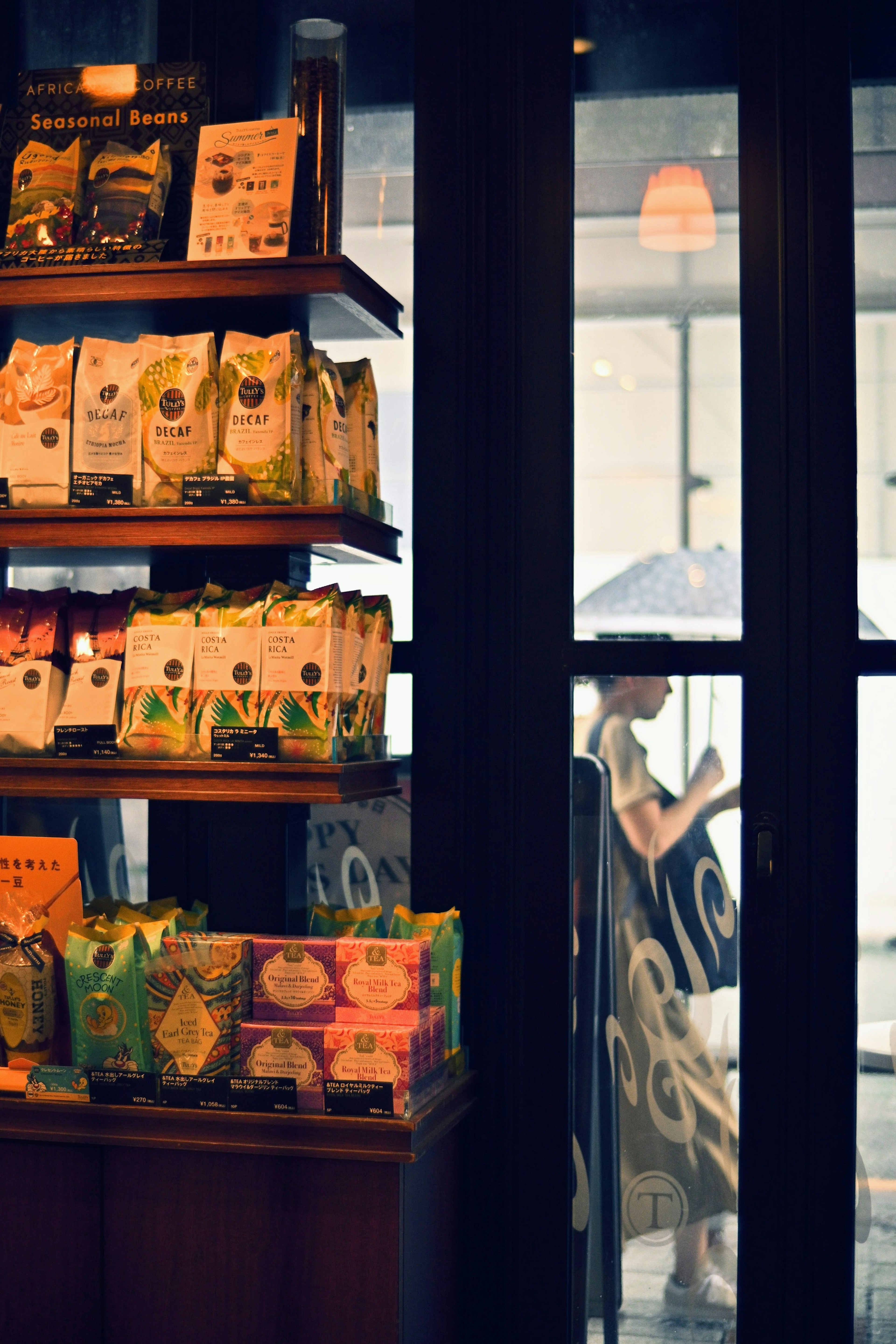 Paquetes de granos de café coloridos en una estantería de una cafetería con una persona usando un teléfono inteligente afuera