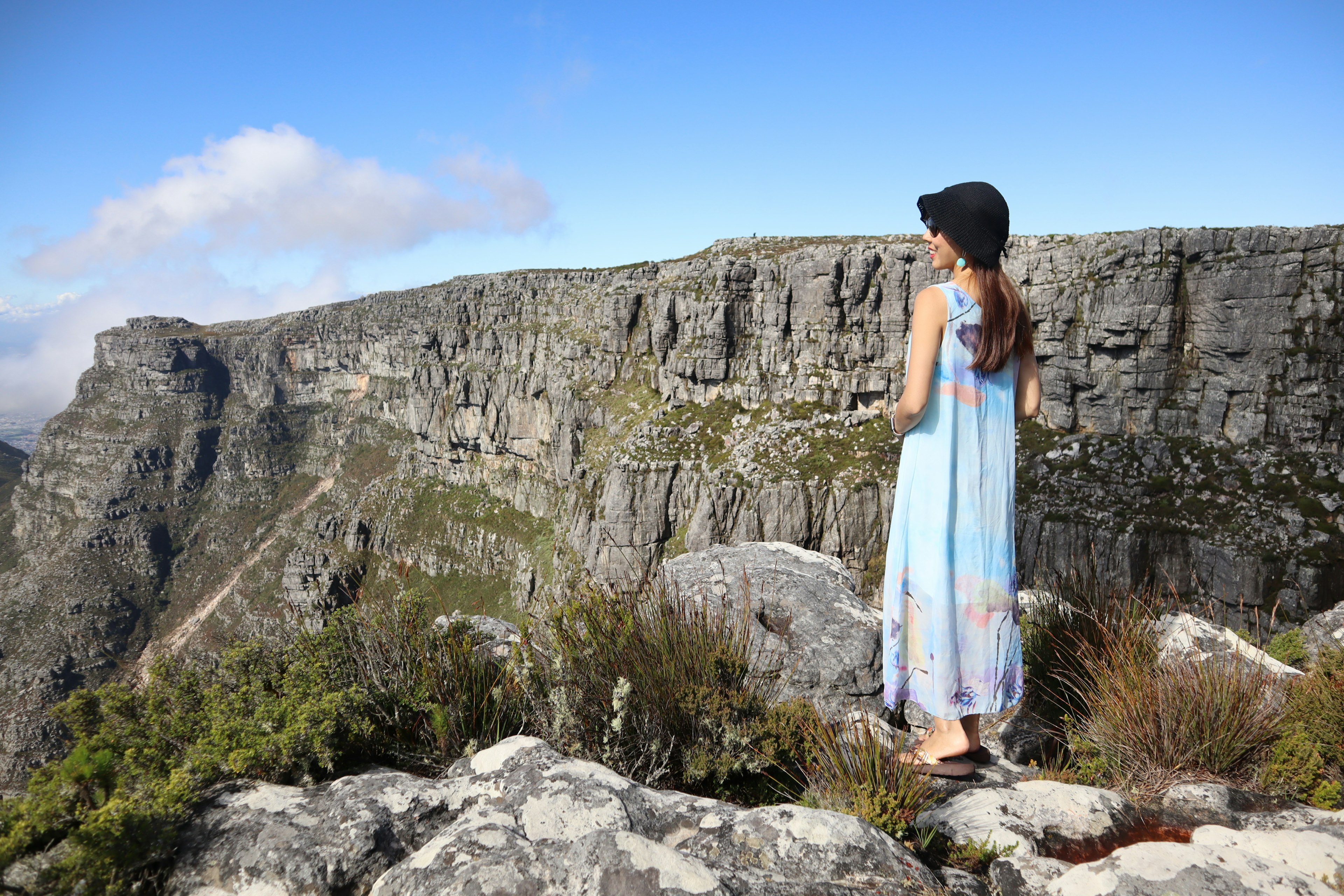 A woman in a blue dress standing on rocks overlooking a majestic cliff under a blue sky