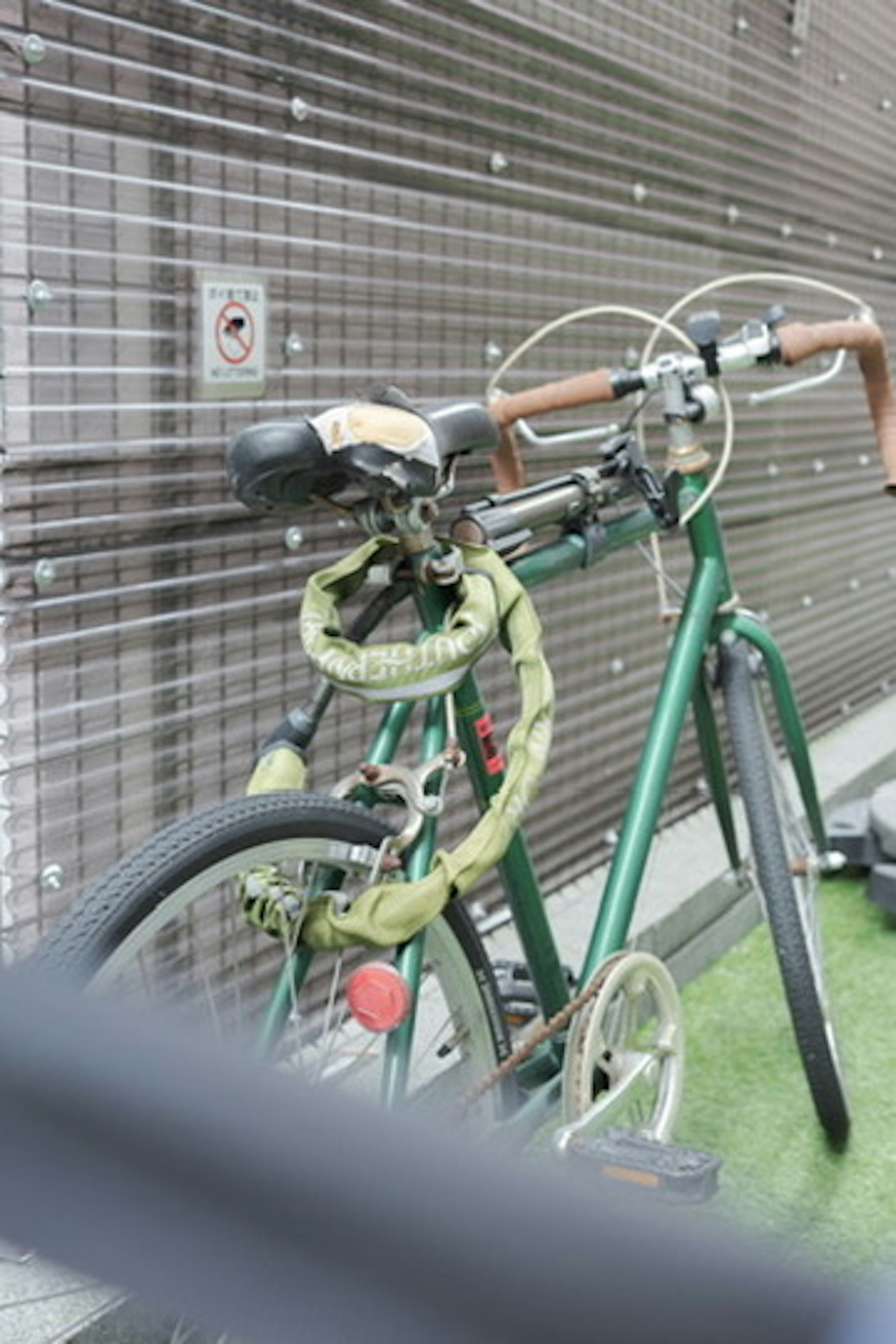 緑色の自転車が壁に固定されている 自転車のハンドルには茶色のグリップがついている 自転車の前輪にはロックがかかっている