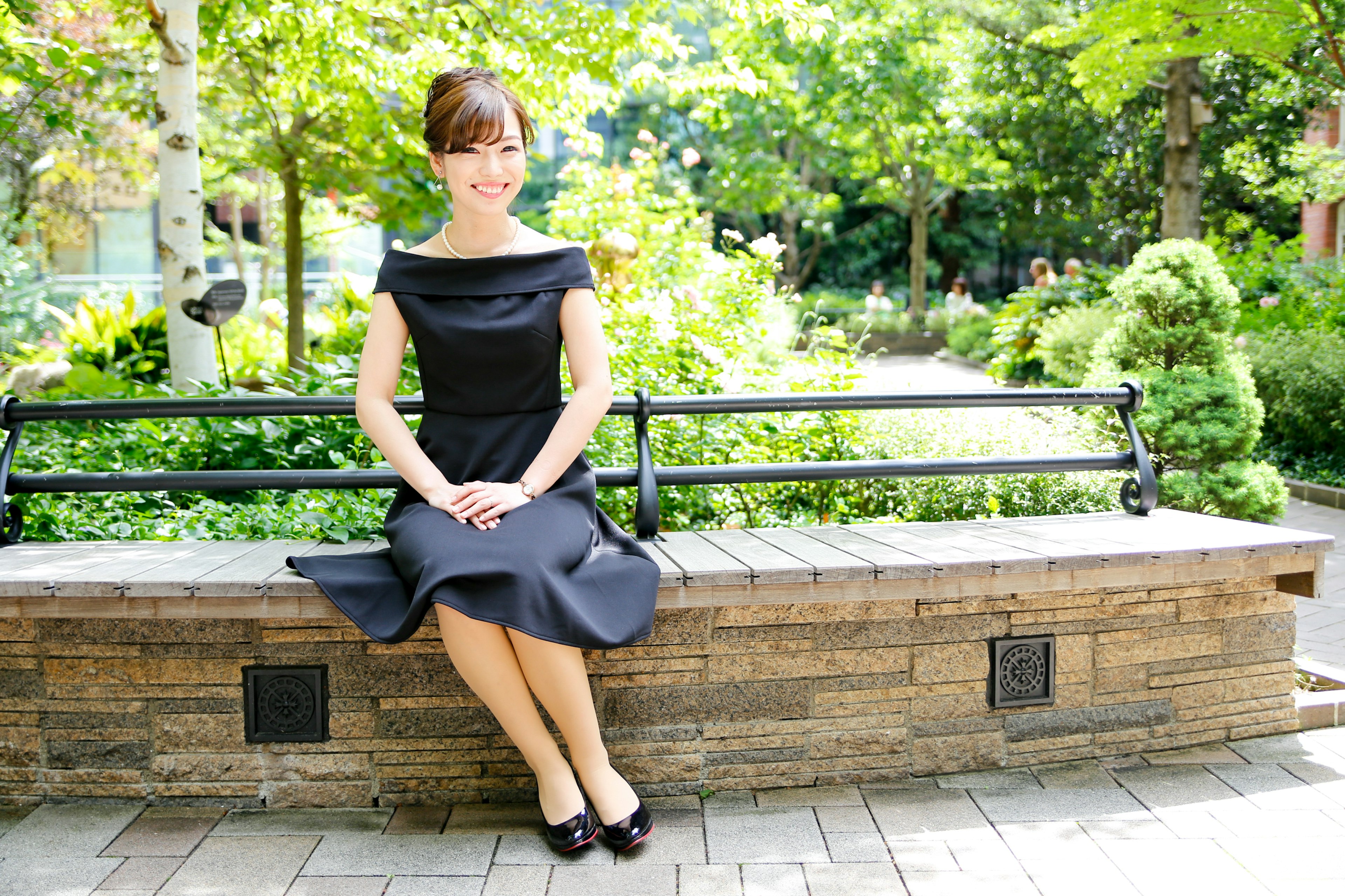 A woman sitting on a park bench wearing a black dress surrounded by lush greenery