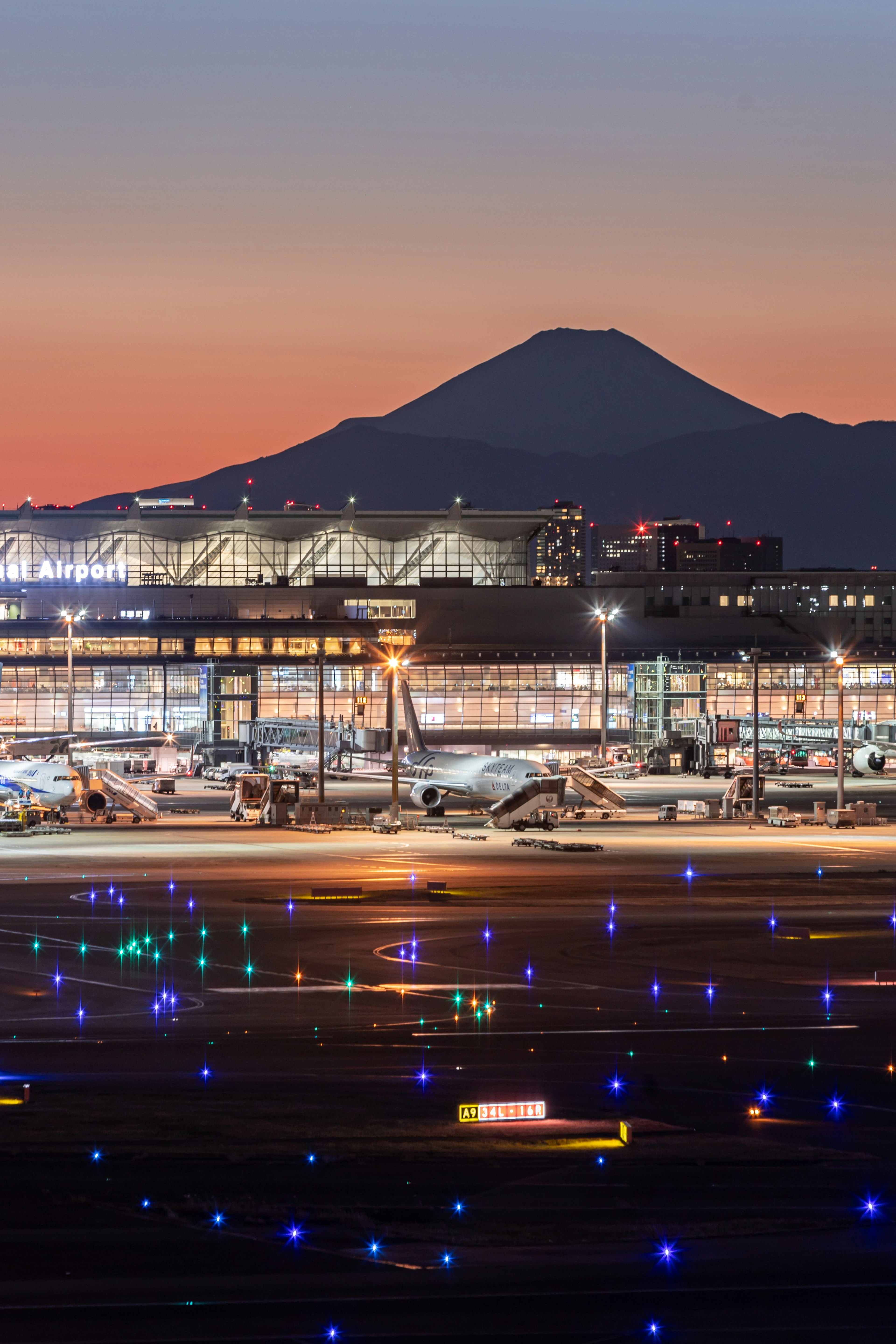 空港の滑走路に青い照明が点灯し背景には富士山が見える夕焼けの風景