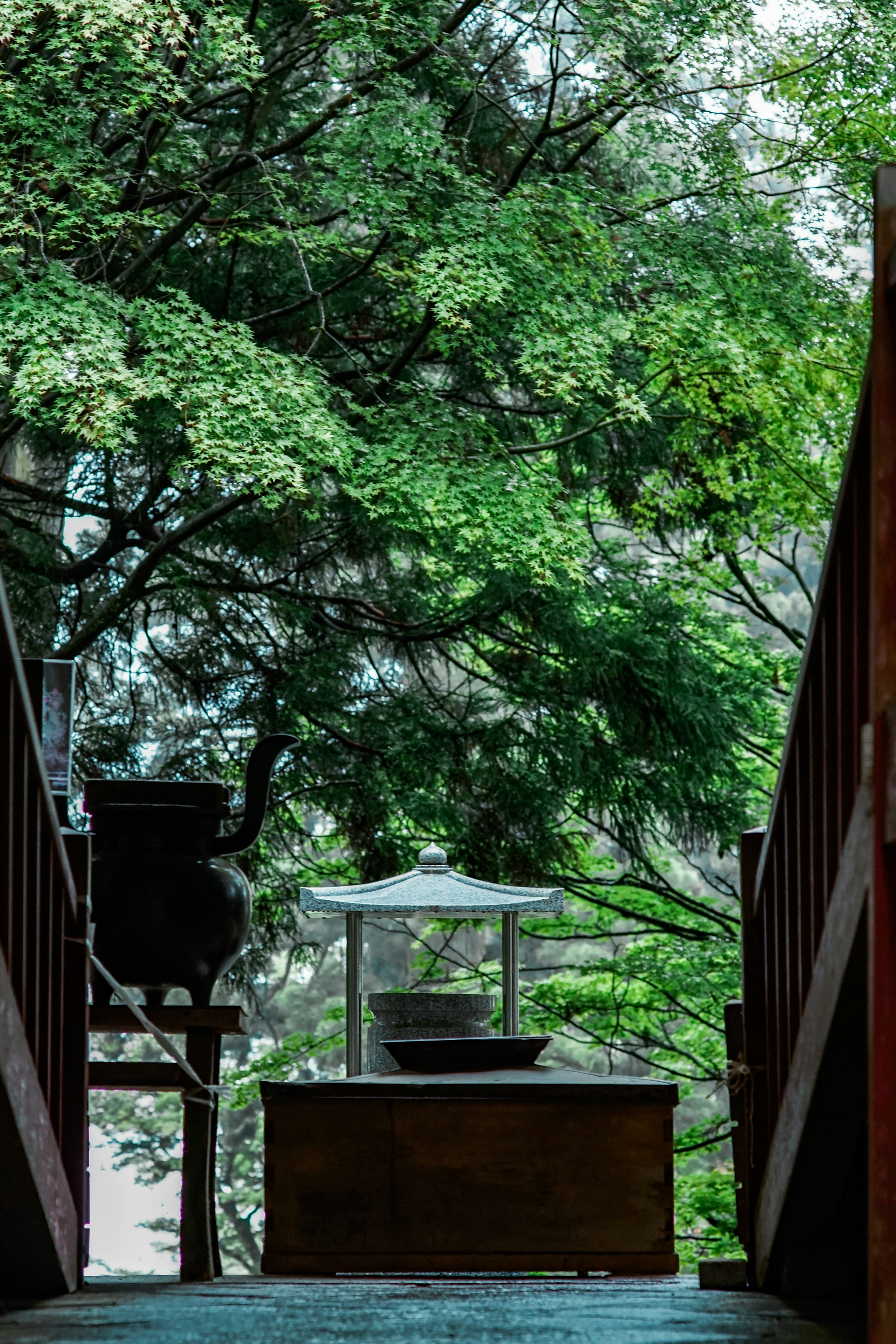 A serene view of a stone lantern and water basin surrounded by lush green trees at the end of a wooden bridge