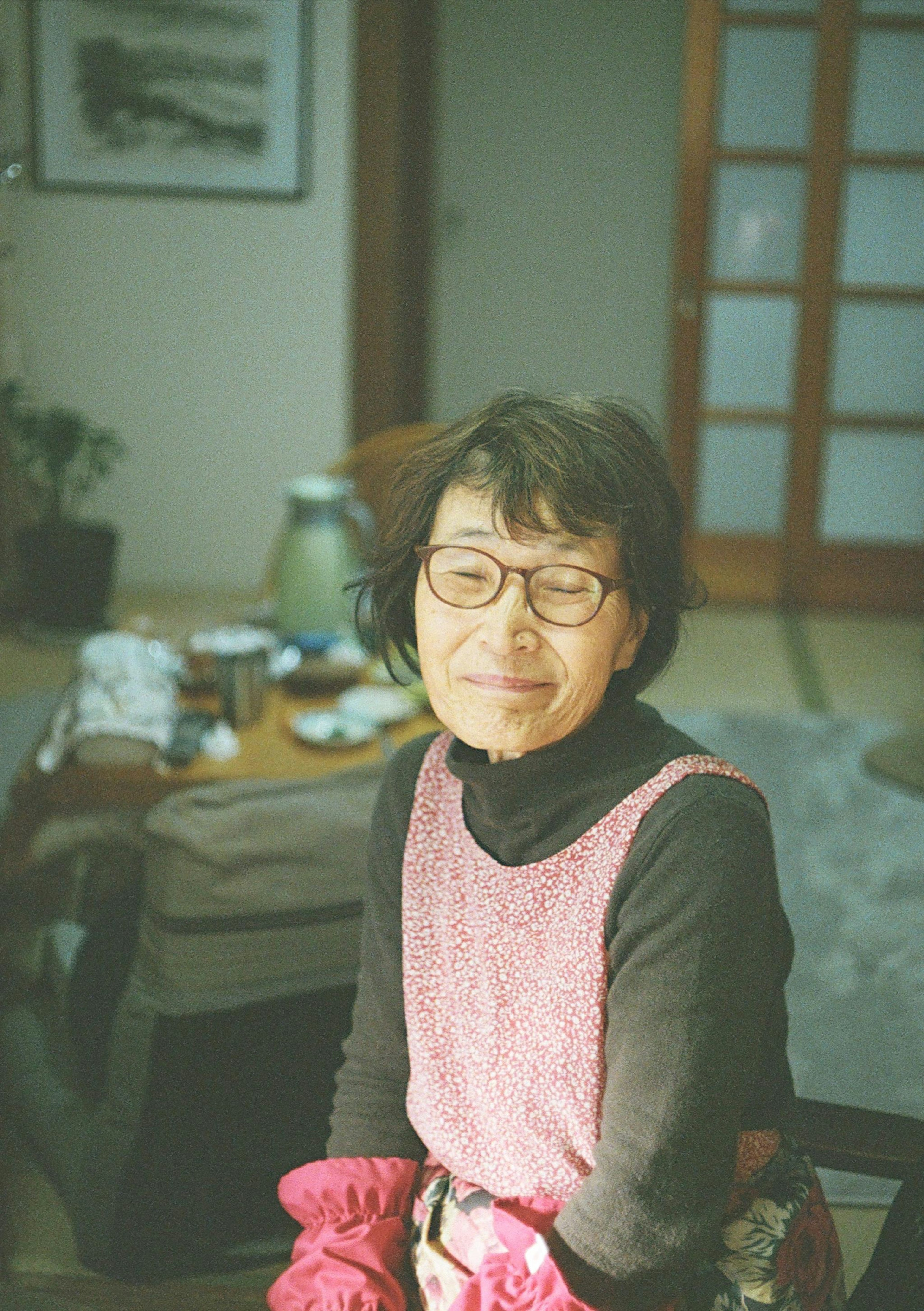 A serene elderly woman wearing a pink apron sitting indoors with a soft expression