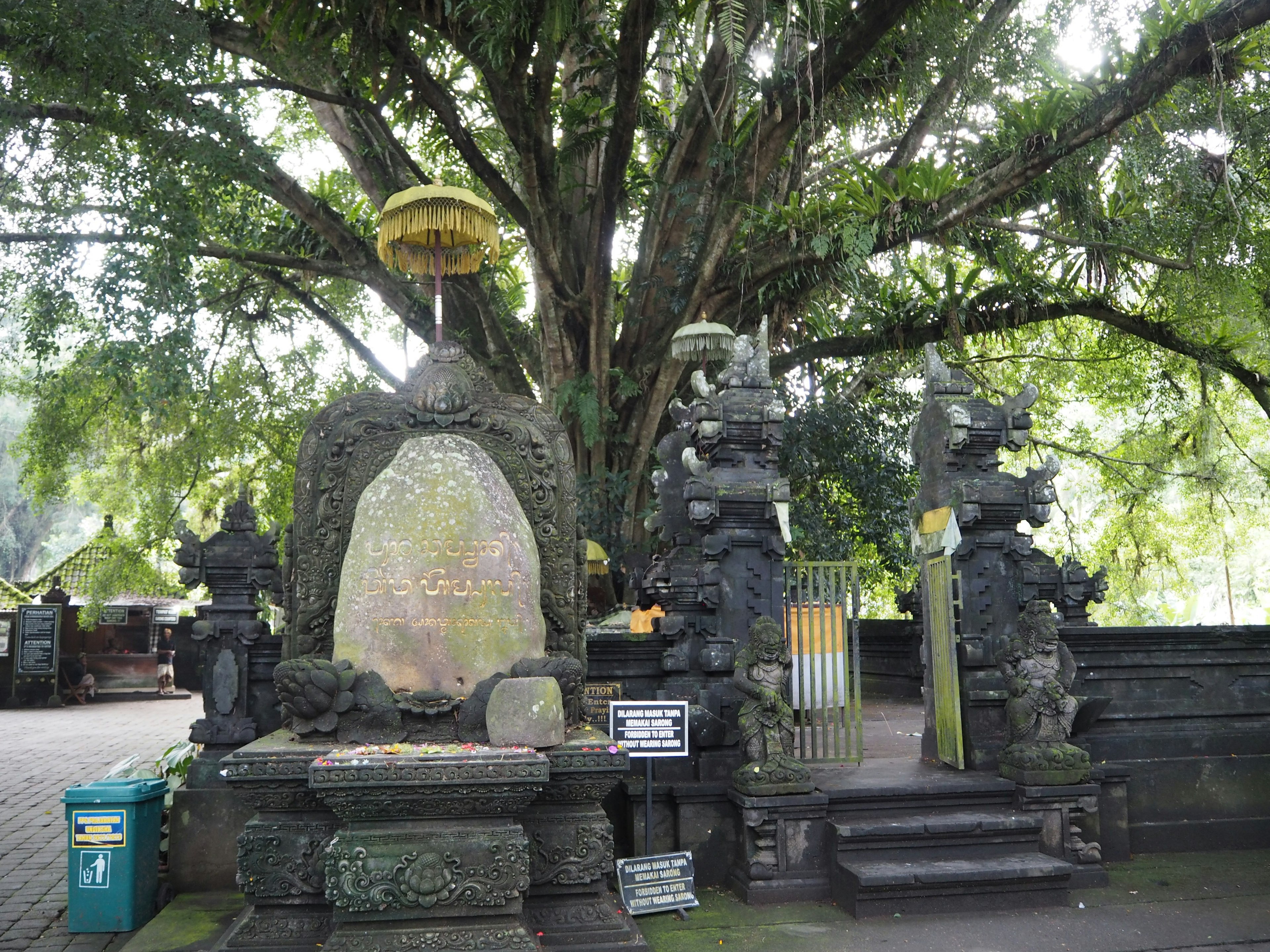 大きな木の下にあるバリ風の石の彫刻と祭壇のある風景