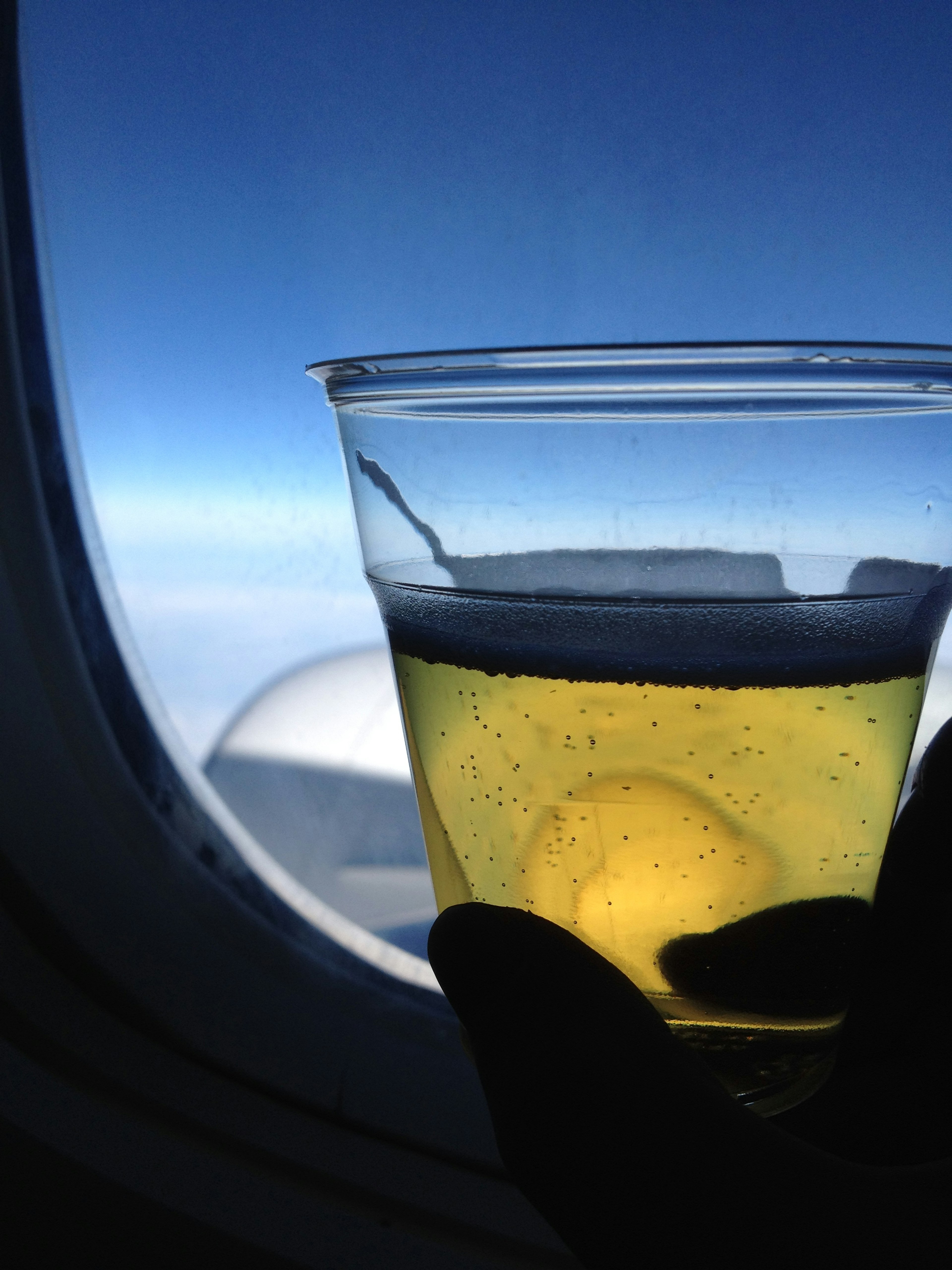 A hand holding a clear cup with a drink against a backdrop of blue sky outside an airplane window