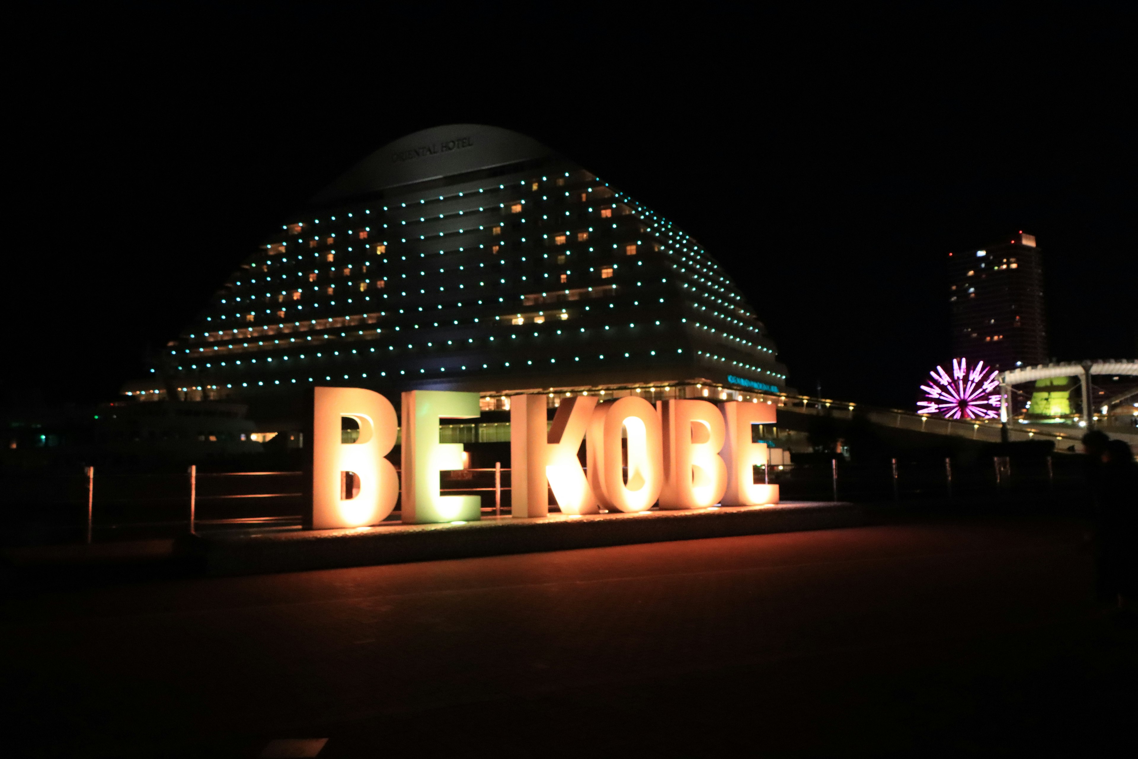 BE KOBE-Schild bei Nacht beleuchtet mit einem kuppelförmigen Gebäude im Hintergrund