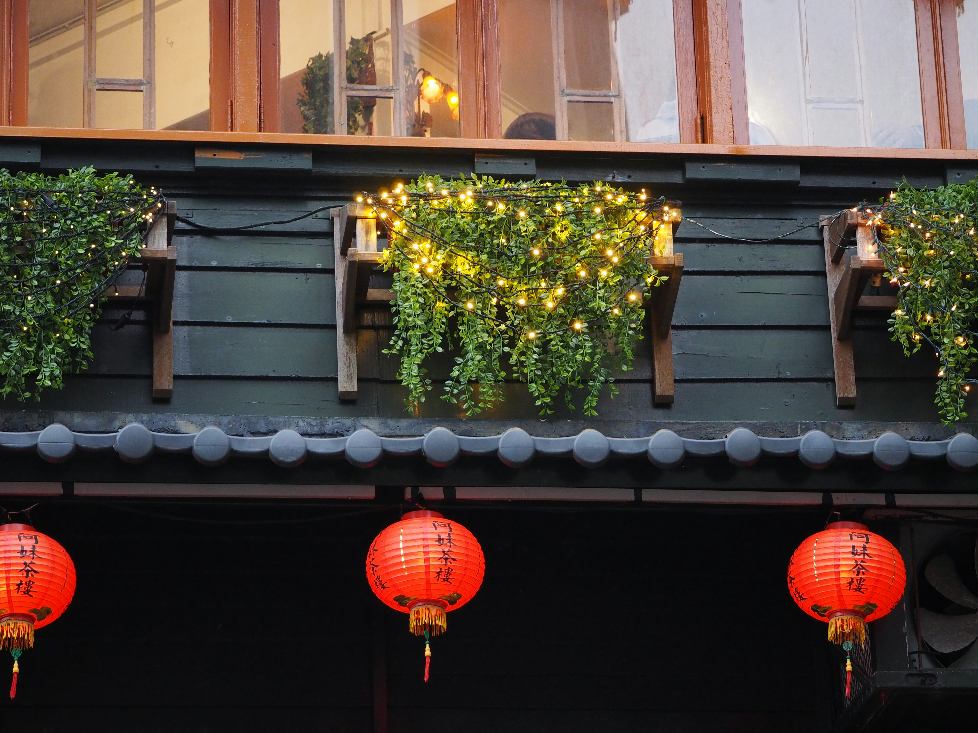 A view of a black wall with green plants and bright lights on the windowsill adorned with red lanterns