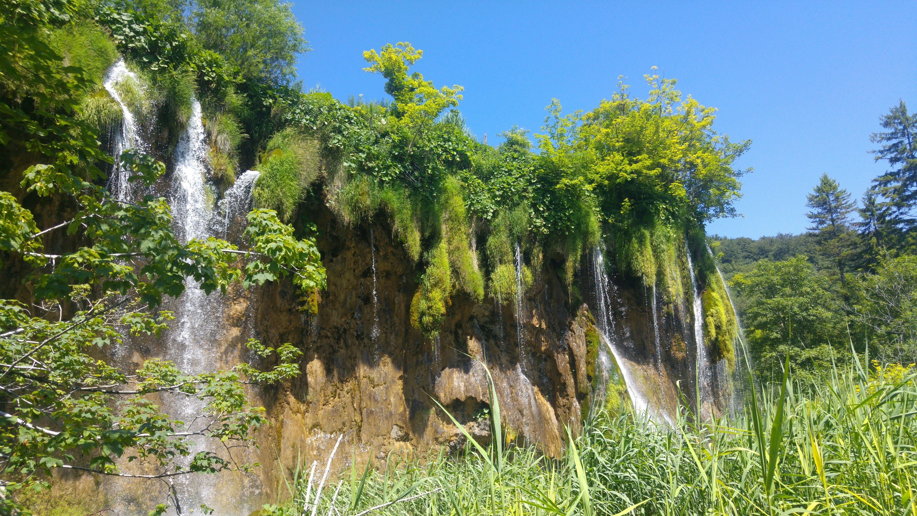 Una vista escénica de cascadas que caen por un acantilado rocoso rodeado de vegetación exuberante bajo un cielo azul despejado