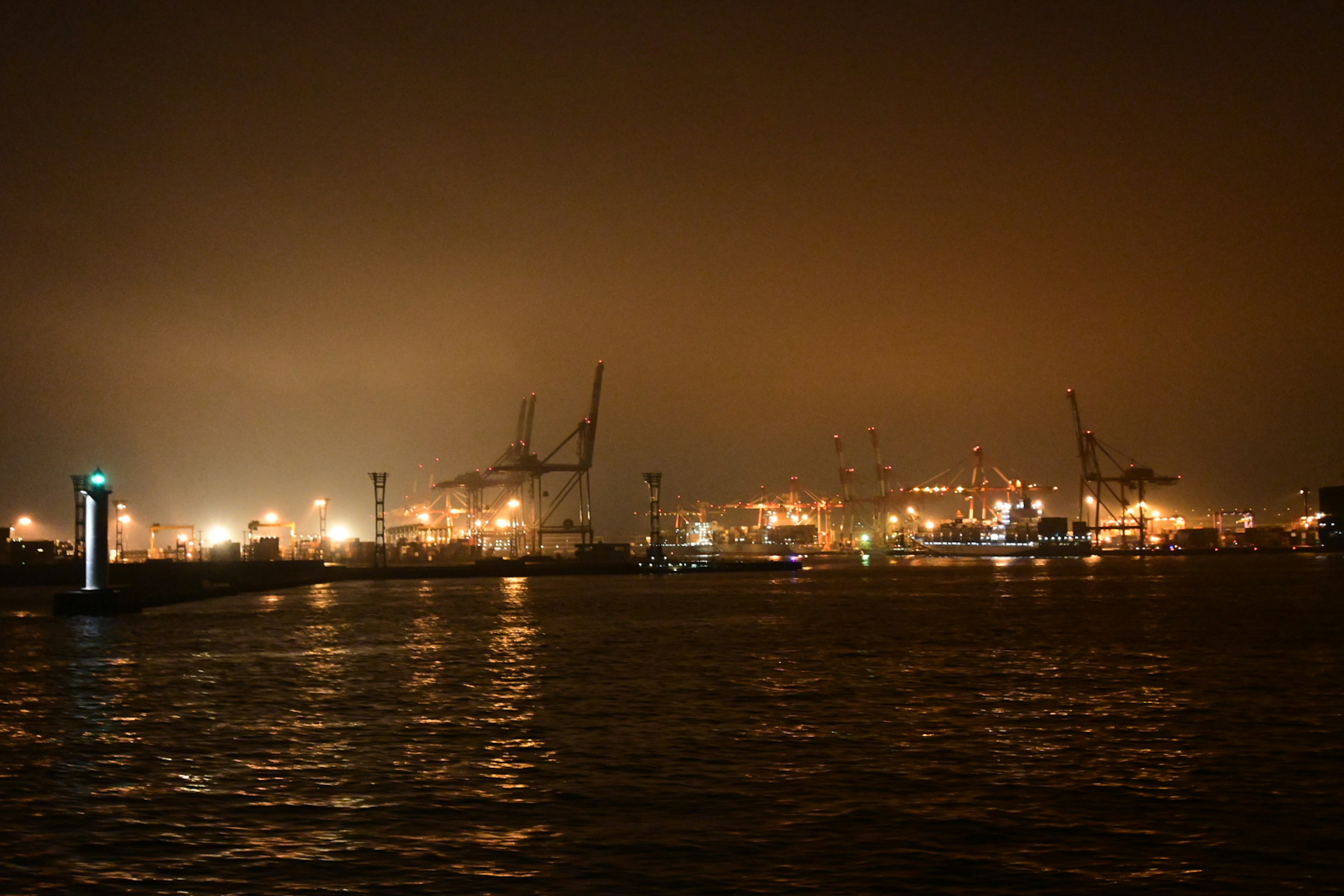 Nighttime harbor scene with shimmering lights on the water and visible cranes