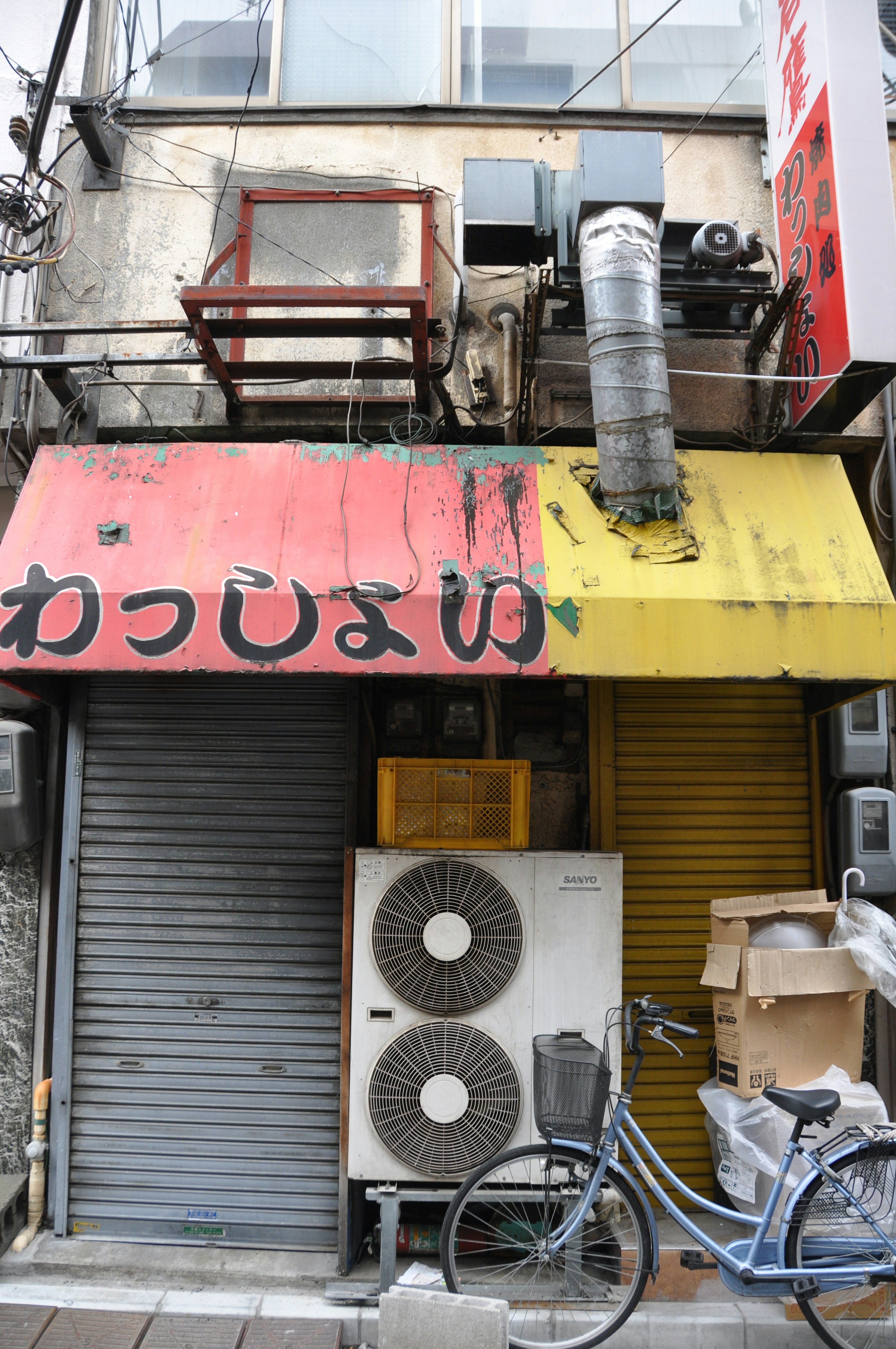 Facade of an old shop with a red and yellow sign and shutter featuring air conditioning units and a bicycle