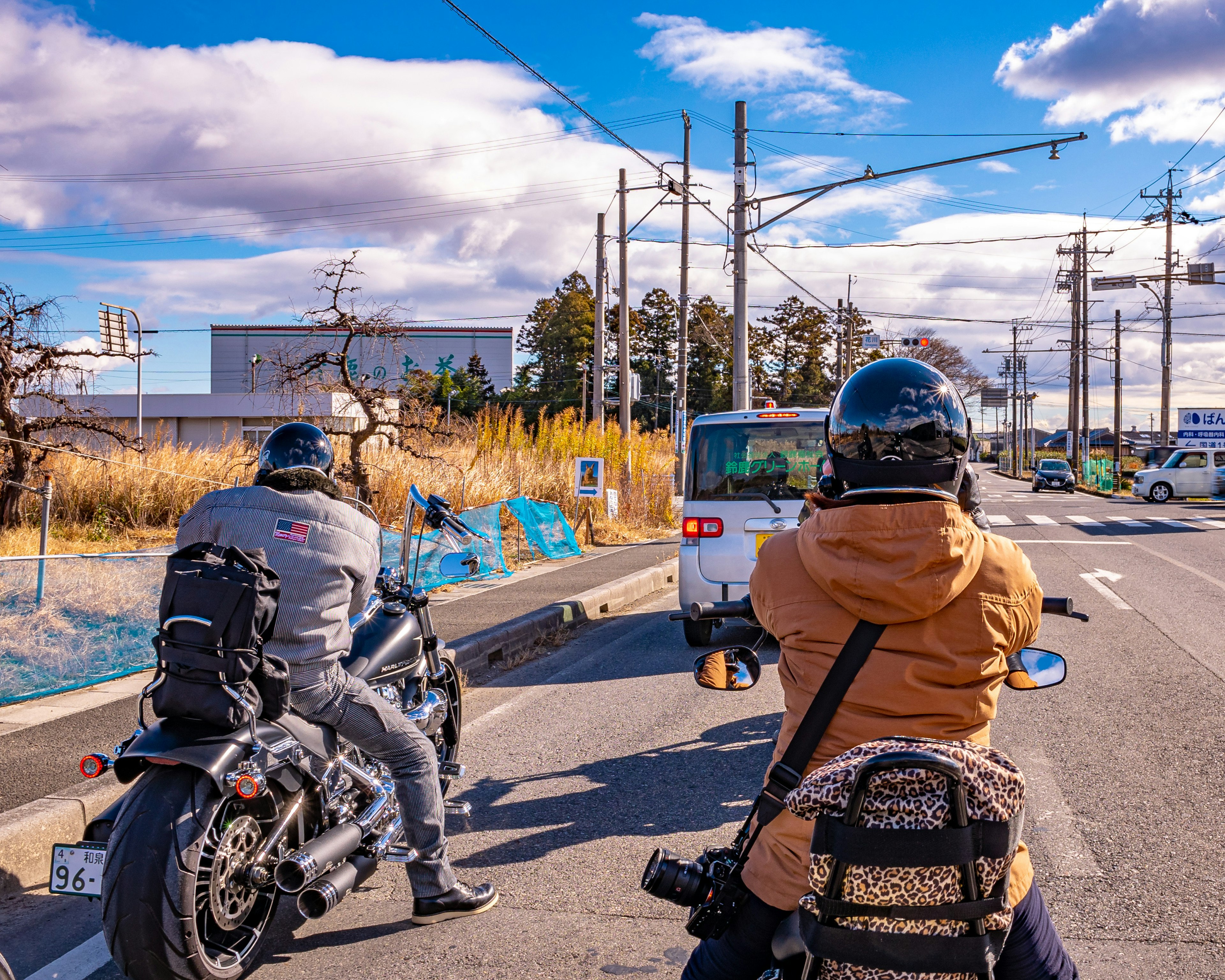 Two riders on motorcycles waiting at a road intersection with a rural background and blue sky