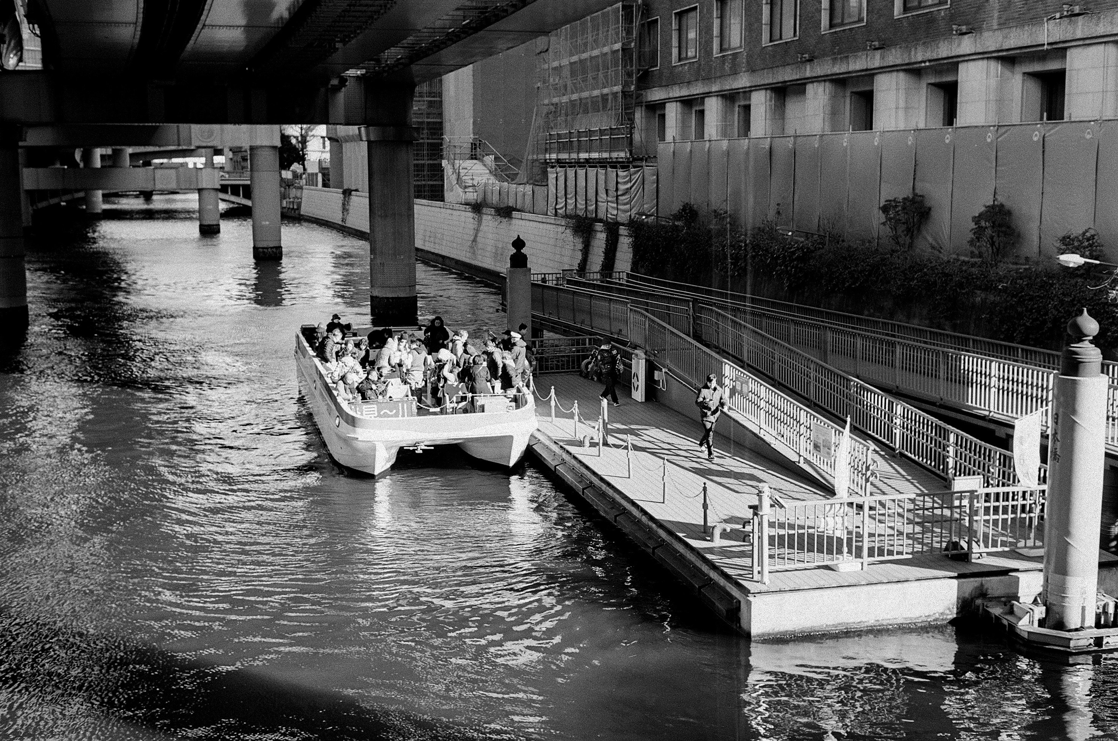 Schwarzweißszene eines Ausflugsboots, das an einem Fluss anlegt, mit Schatten von Hochhäusern