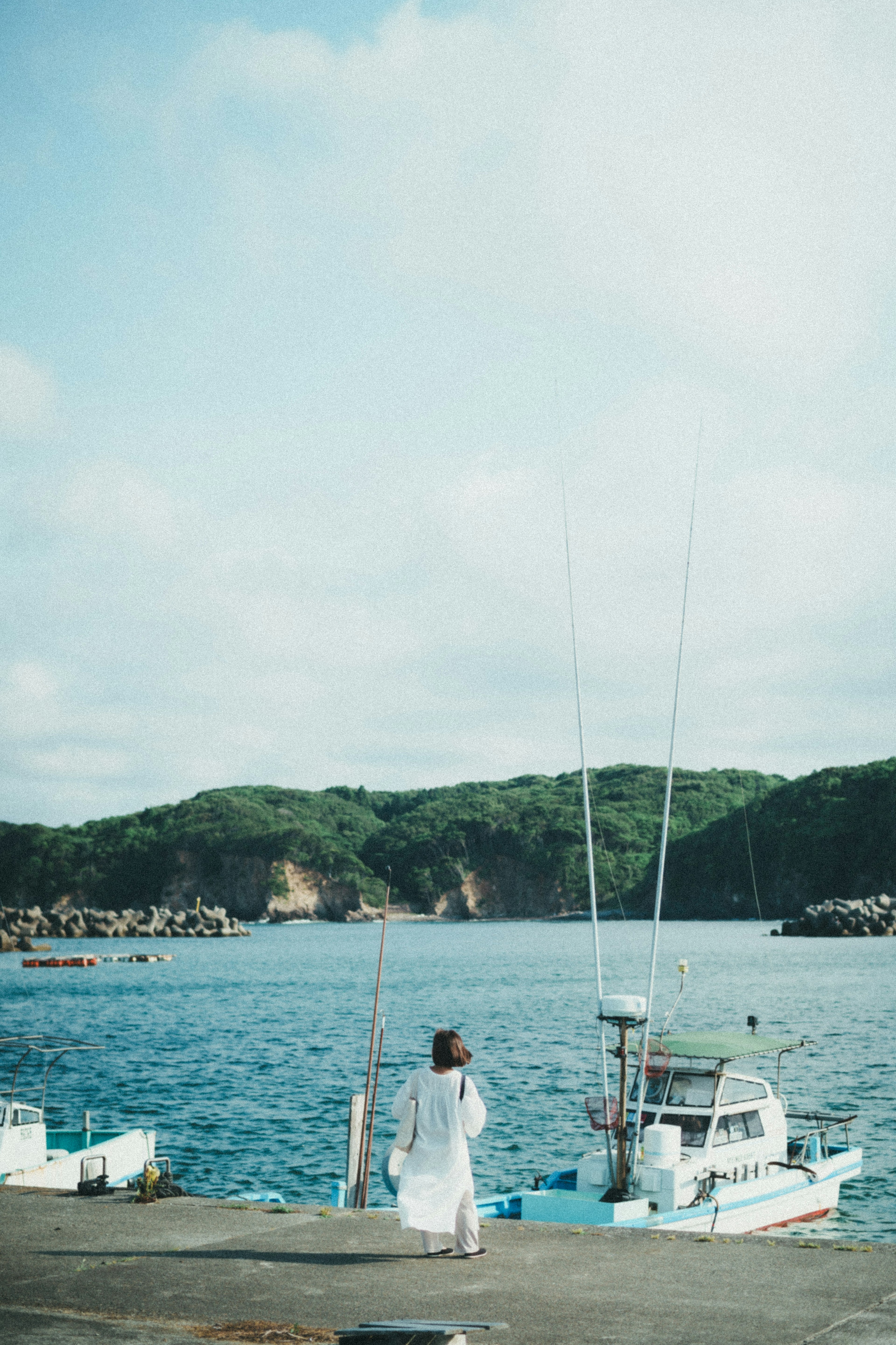 Una mujer con un vestido blanco caminando hacia el mar con barcos y colinas al fondo