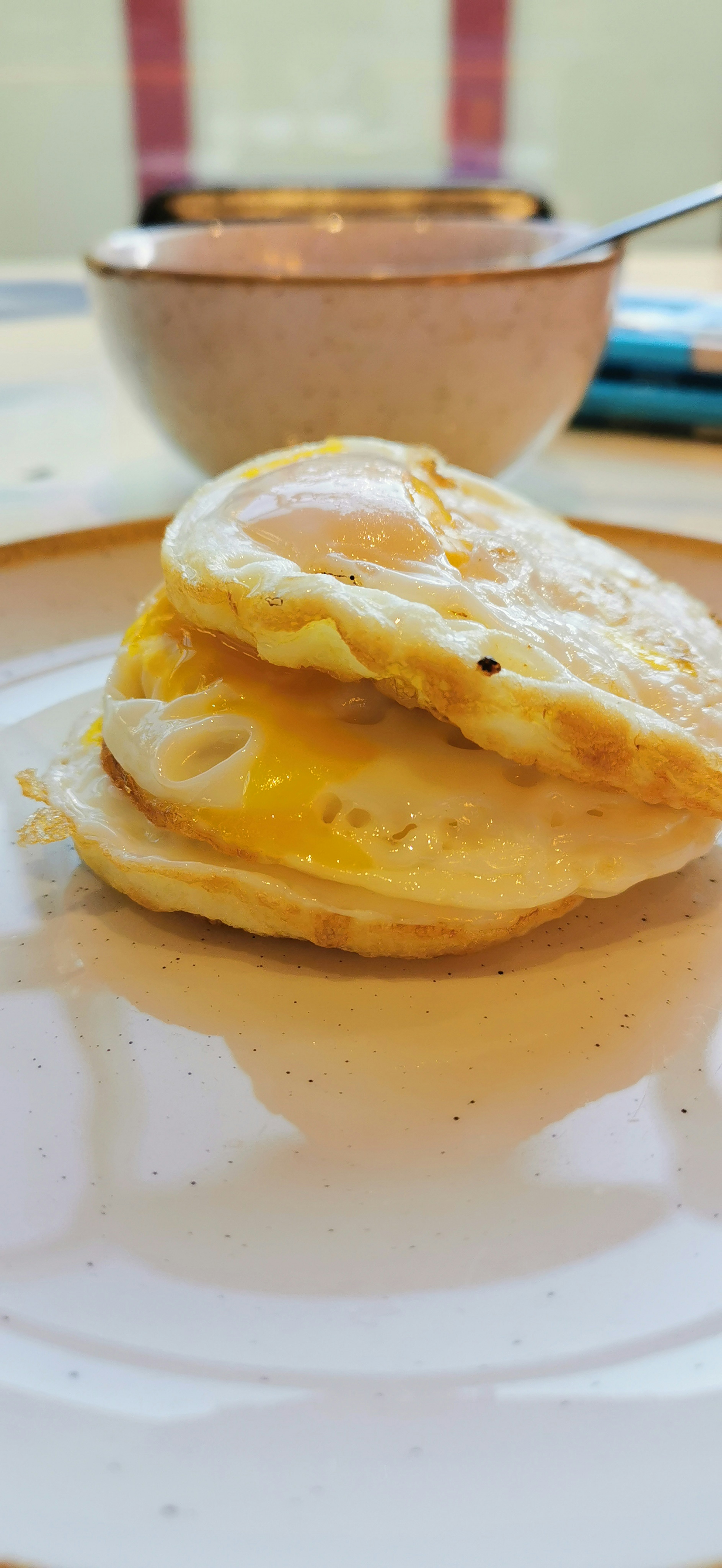 Dos huevos fritos apilados en un plato con un tazón de sopa al fondo