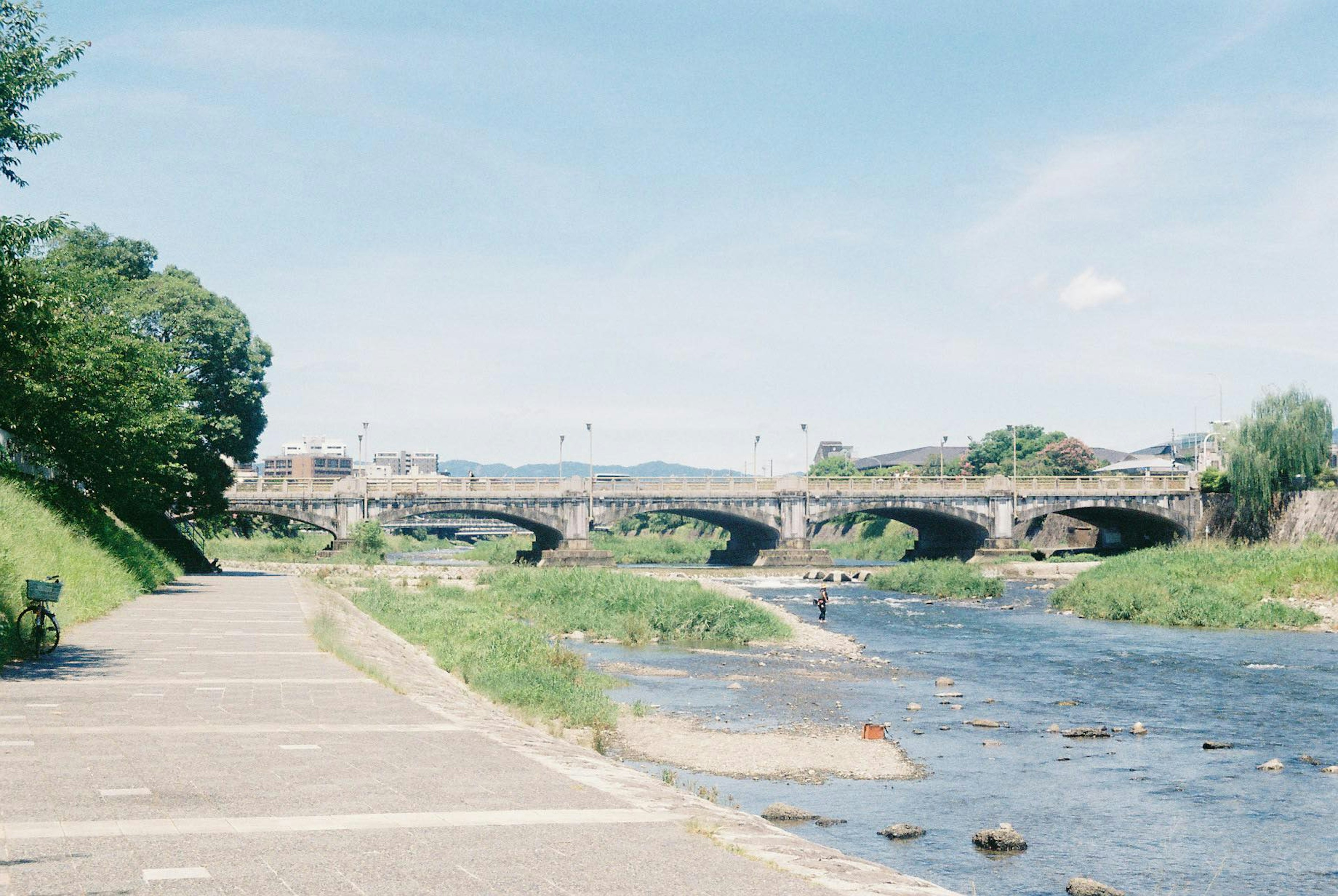 Scenic riverside walkway with a bridge under a clear blue sky and lush greenery