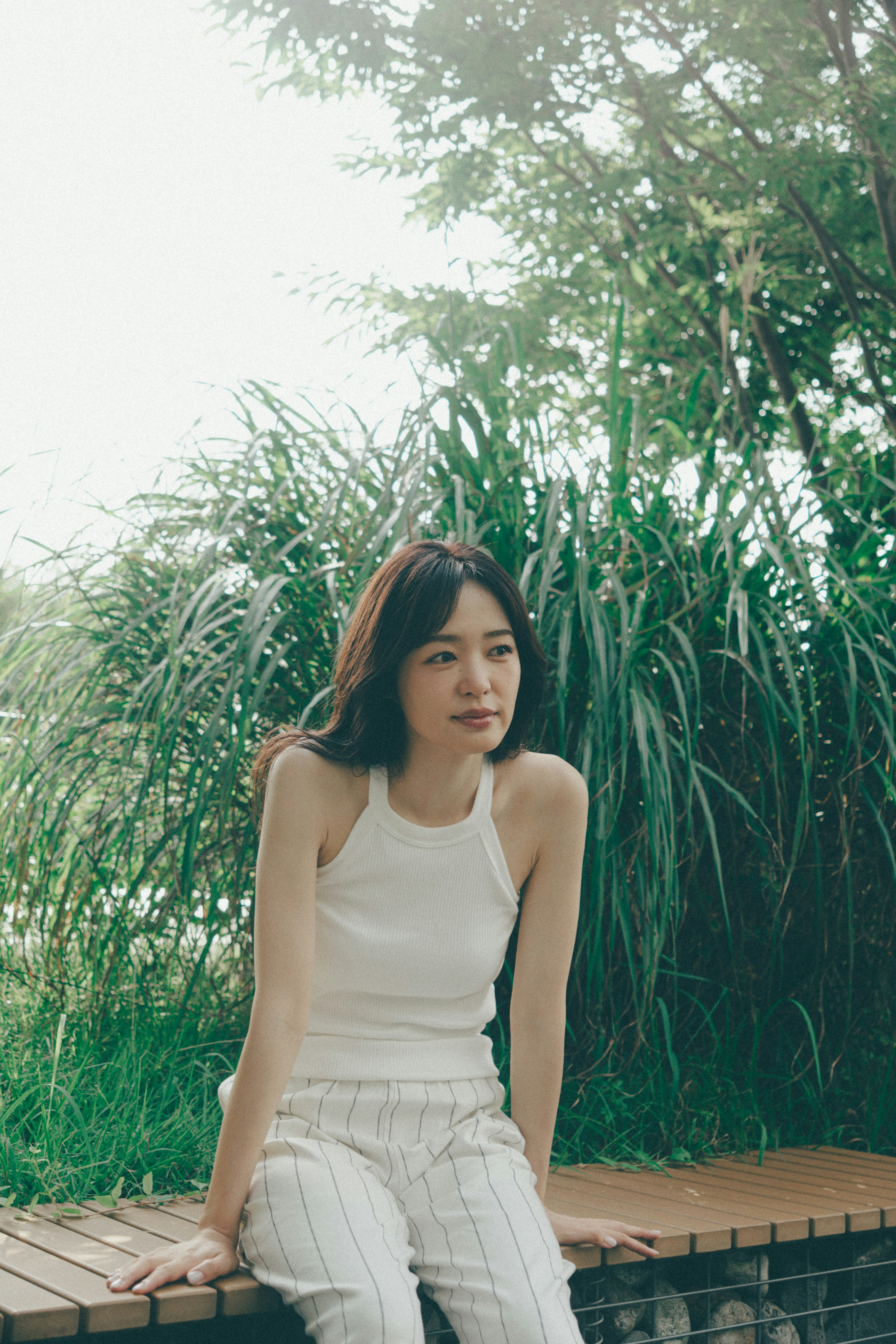 A woman in a white tank top sitting in front of green foliage