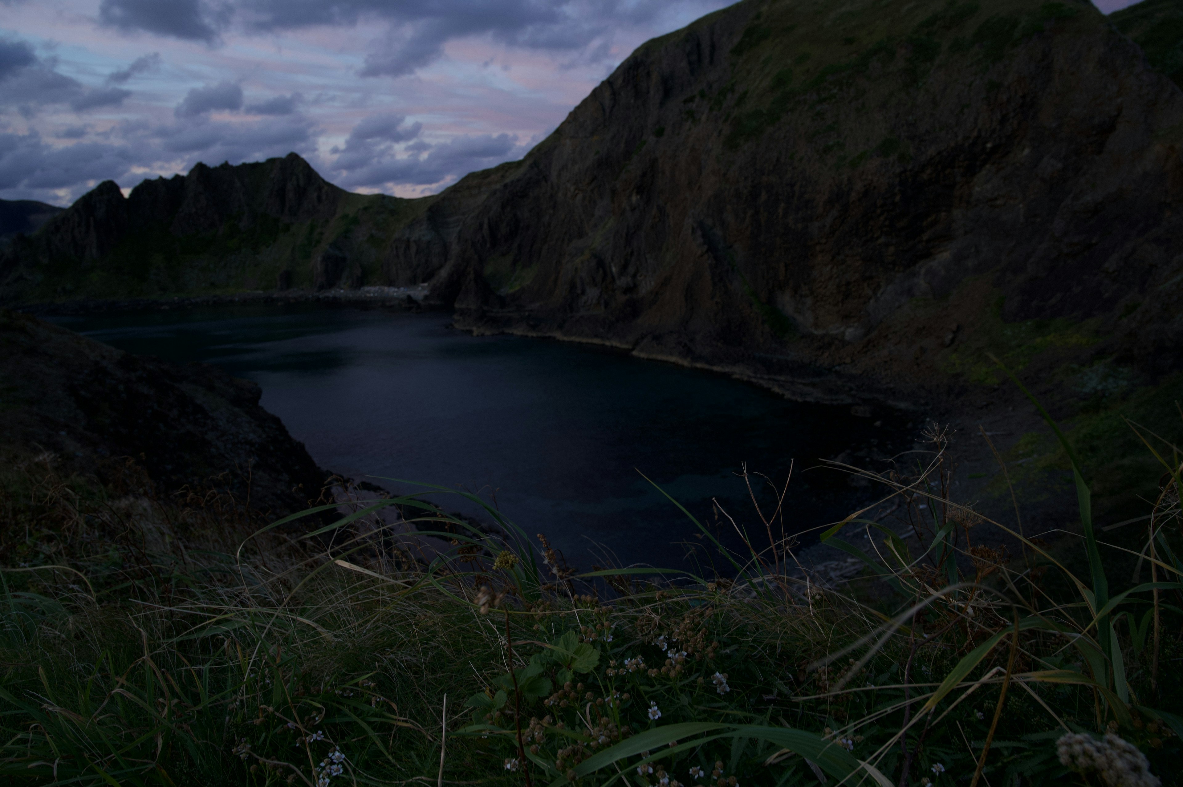 Lago oscuro rodeado de montañas escarpadas al anochecer con nubes dispersas