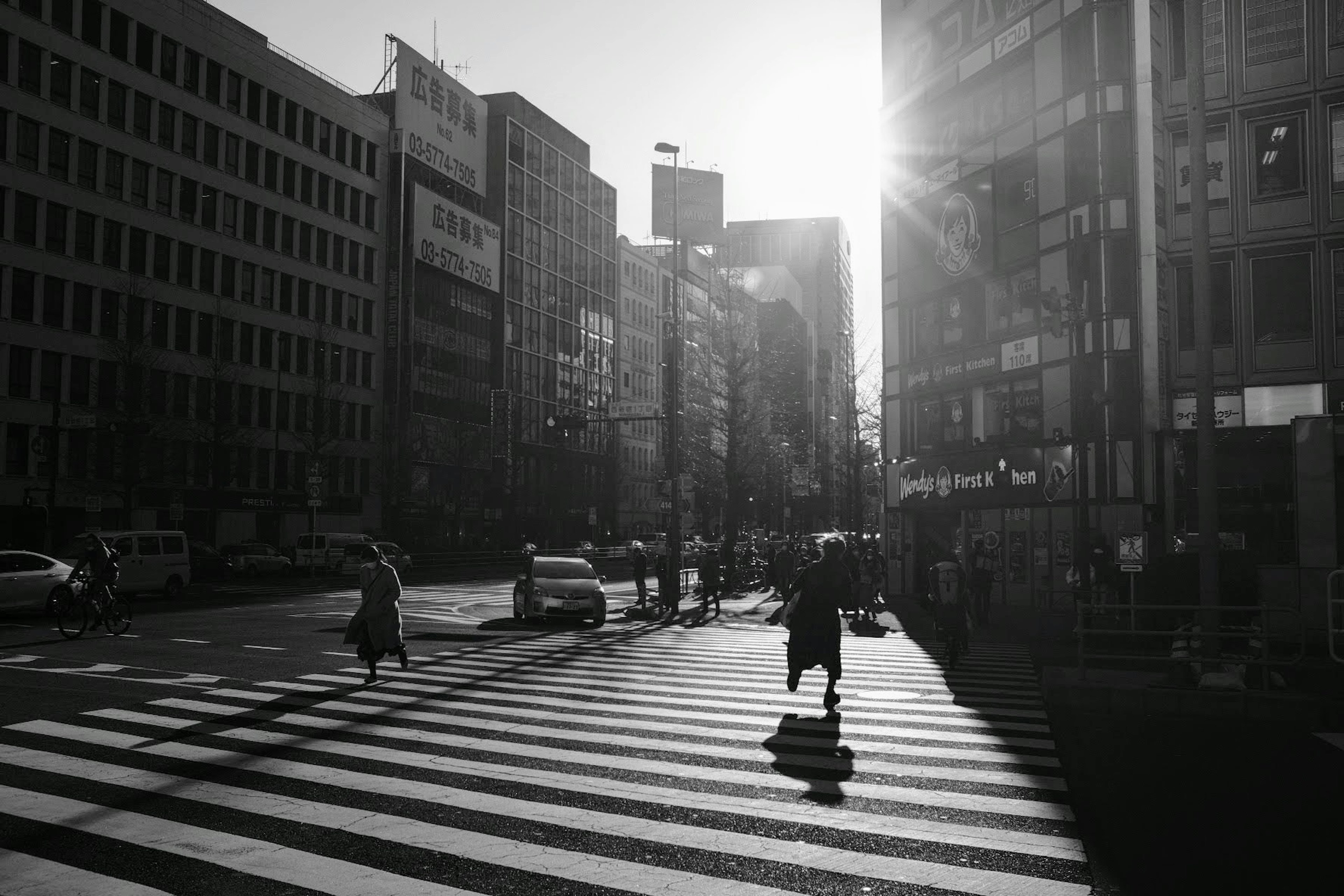 白黒の街角で横断歩道を渡る人々と背後に輝く太陽