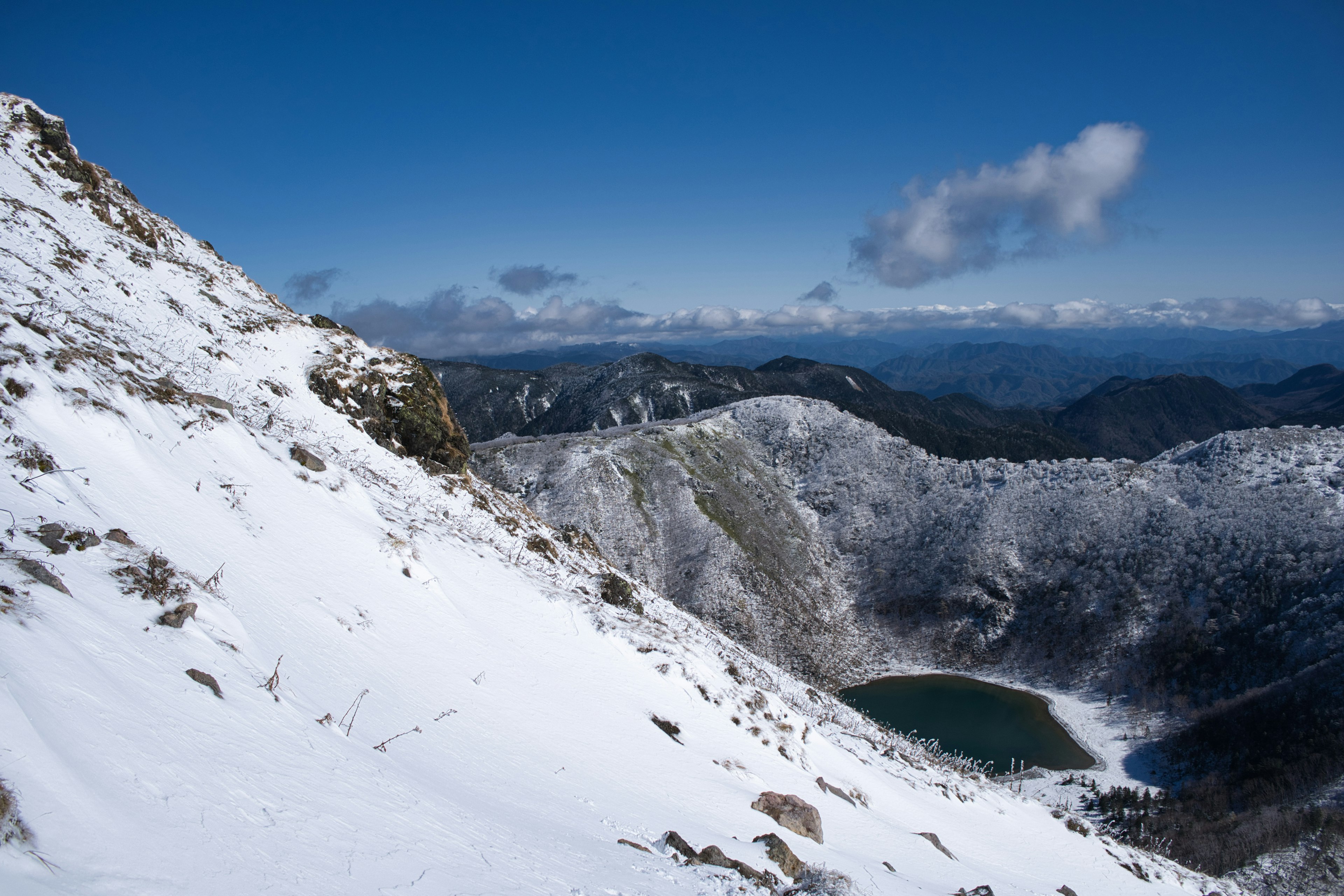 Pendio di montagna coperto di neve con un lago blu sullo sfondo e una vasta catena montuosa