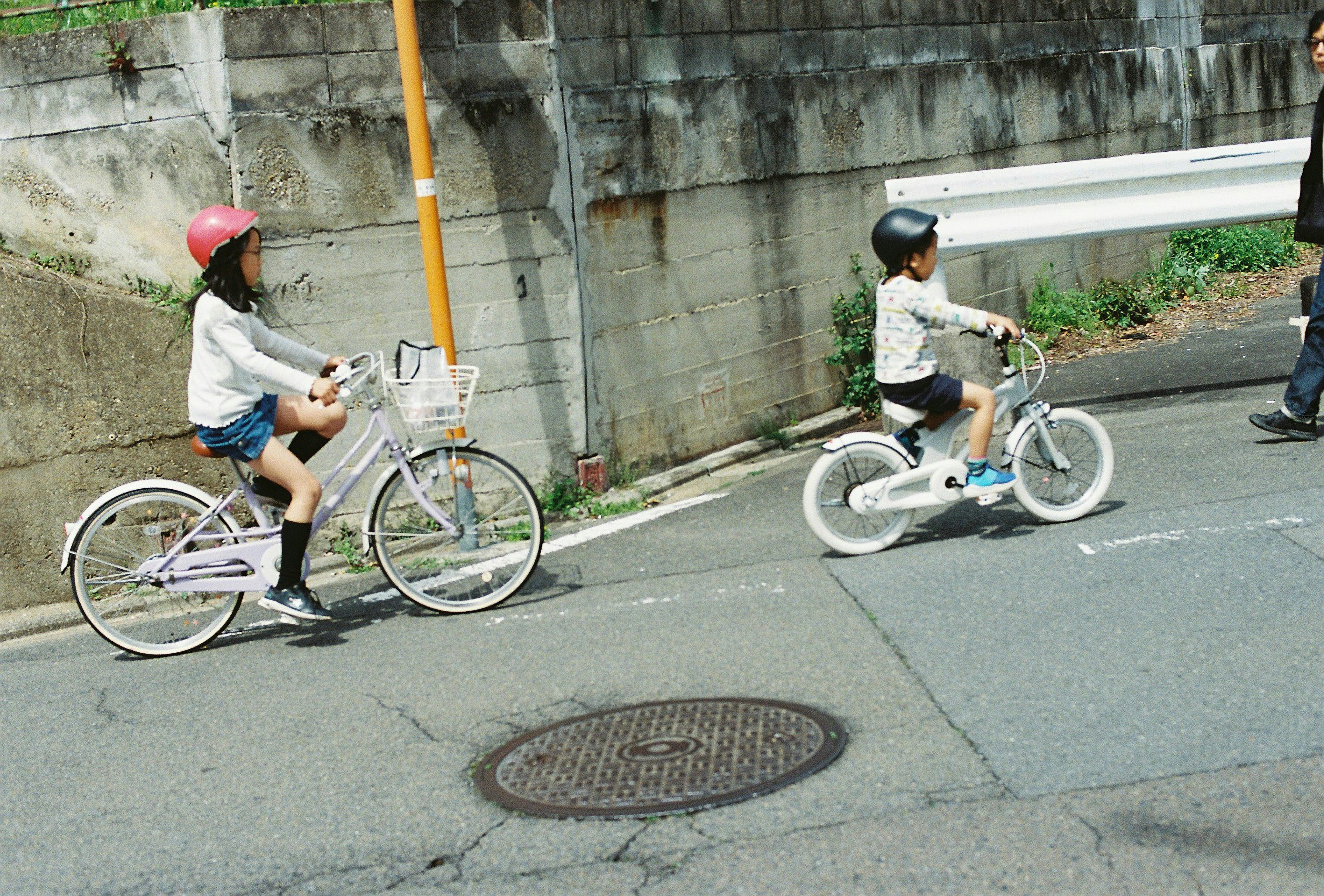 Deux enfants faisant du vélo Une fille porte un casque rose et roule sur un vélo blanc Un garçon porte un casque noir et roule sur un vélo plus petit L'arrière-plan présente un mur en béton