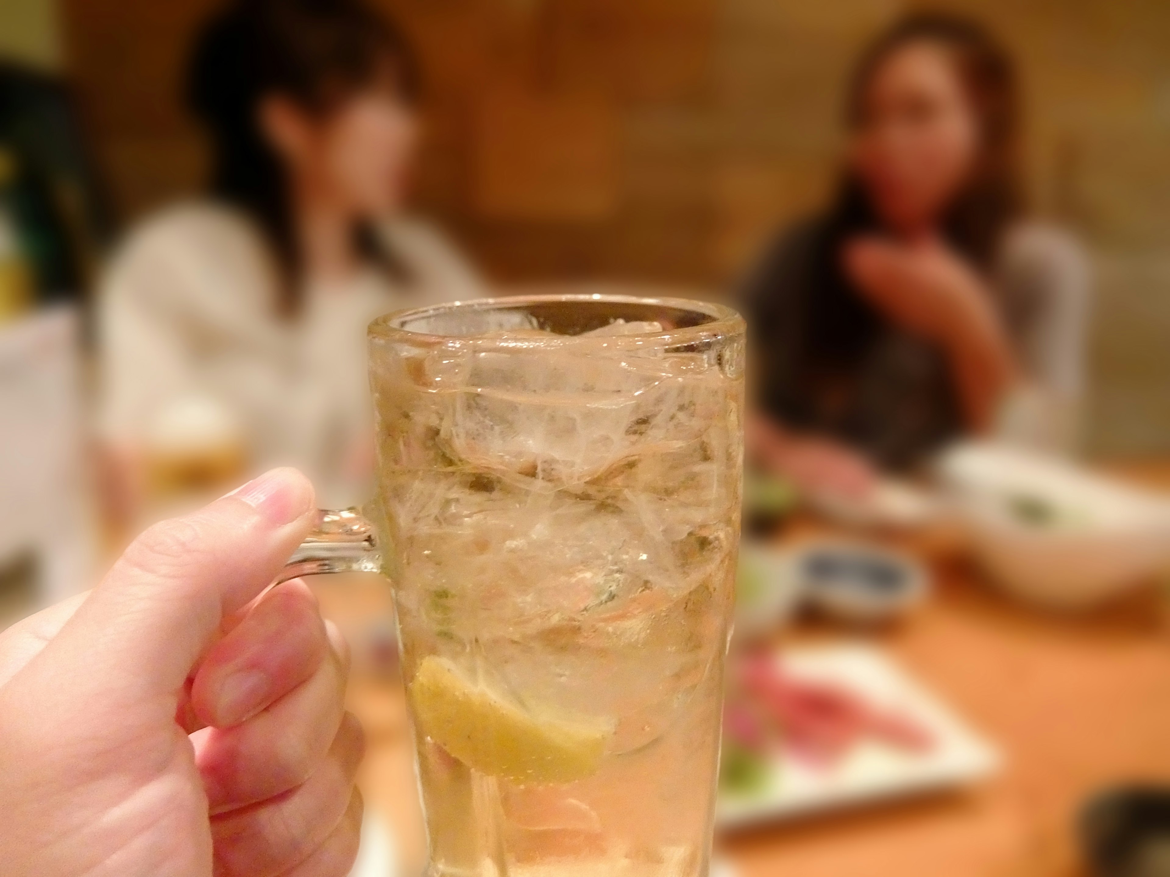 Mano sosteniendo un vaso con hielo y una rodaja de limón y dos mujeres al fondo