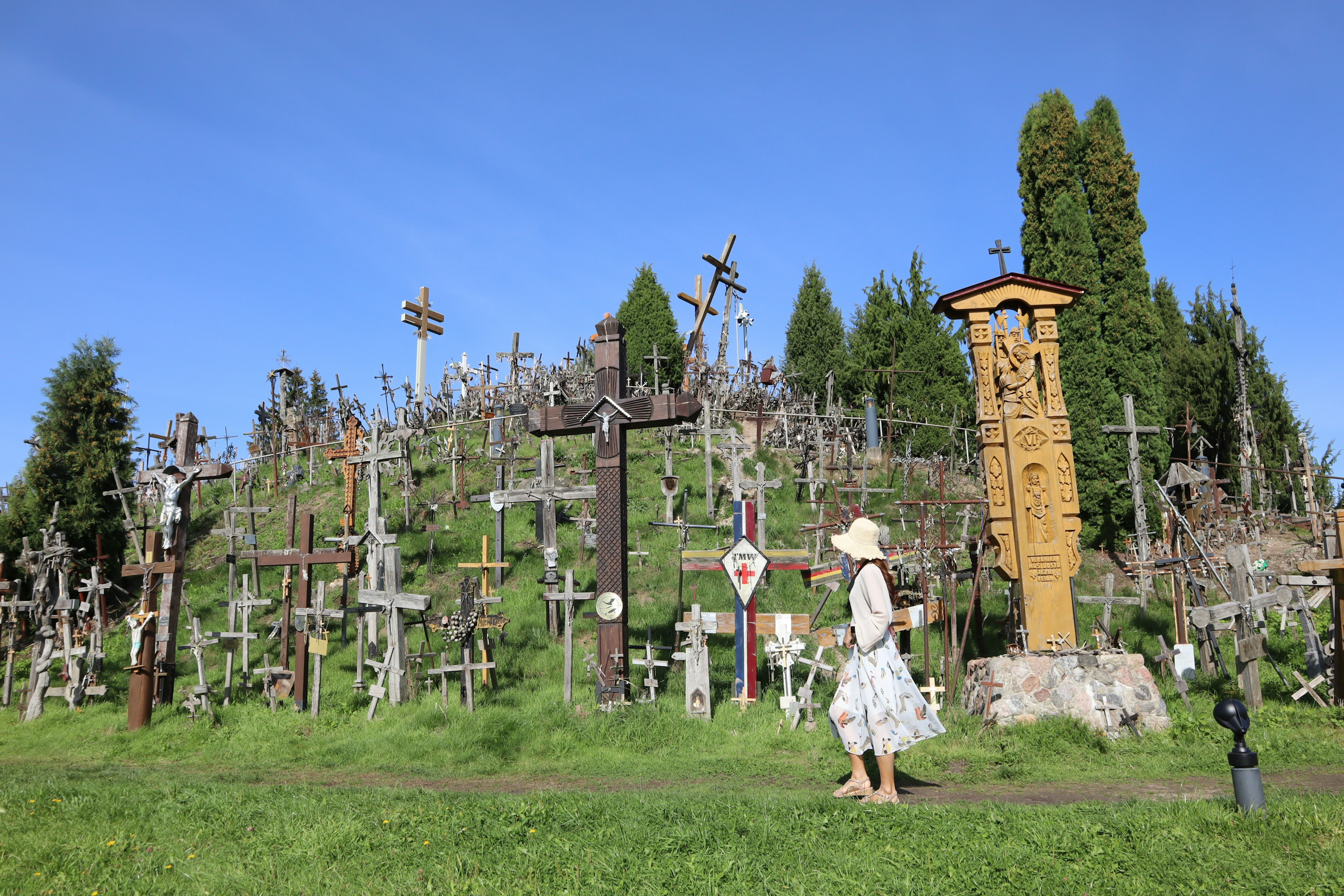 Eine Frau in einem weißen Kleid und Hut, die durch einen Hügel von Kreuzen geht üppiges grünes Gras und blauer Himmel im Hintergrund