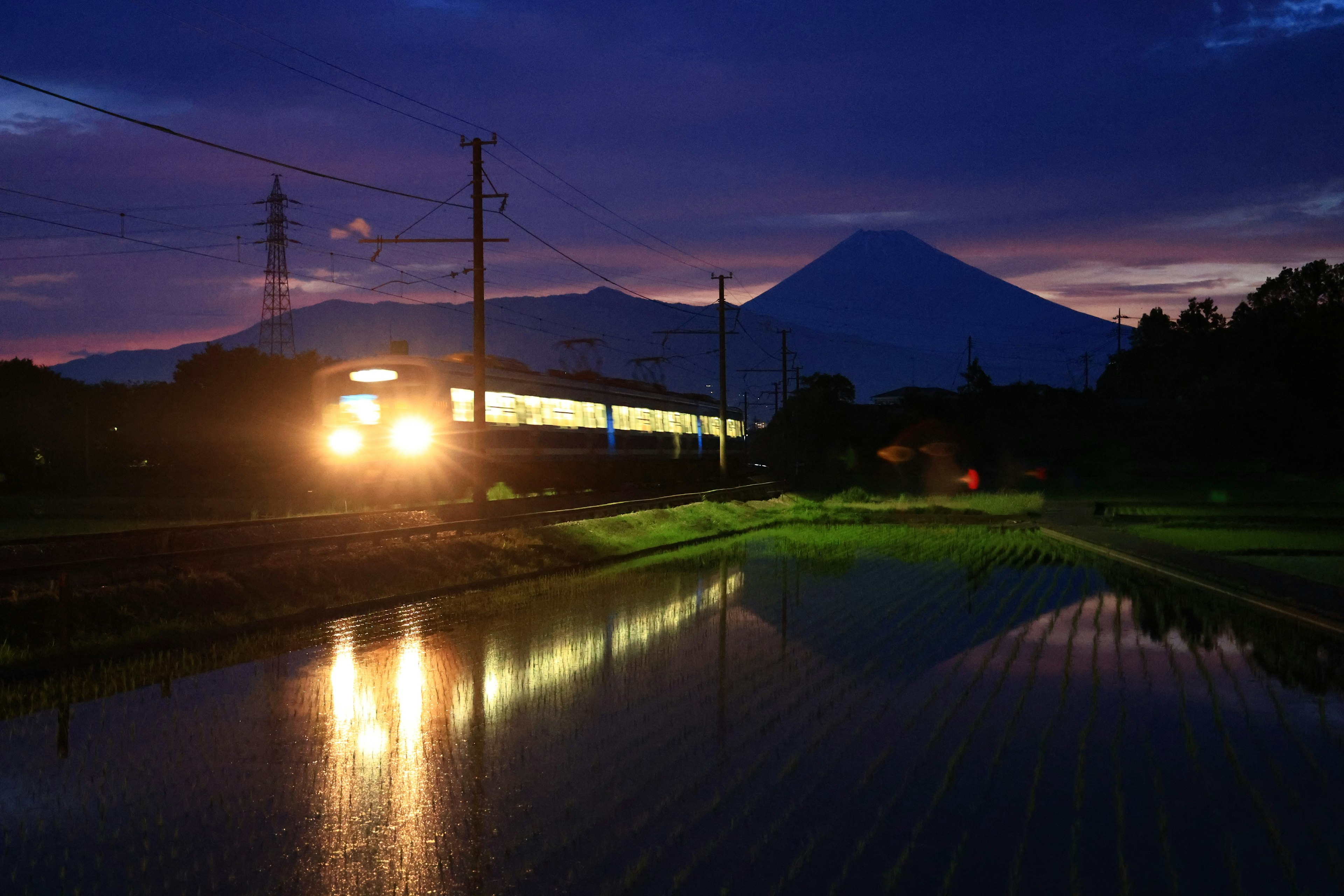 夕暮れ時に富士山を背景に走る列車と水田の反射